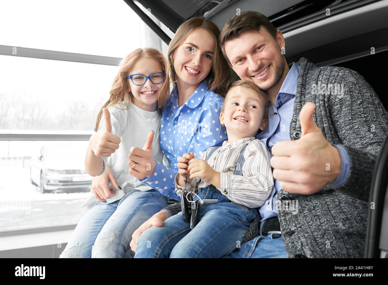 La famiglia felice in seduta nuove e confortevoli auto in auto salon, guardando la telecamera, sorridente e mostra segno ok. Incantevoli i genitori con bambini acquisto di veicolo in showroom. Concetto di automobile l'acquisto. Foto Stock