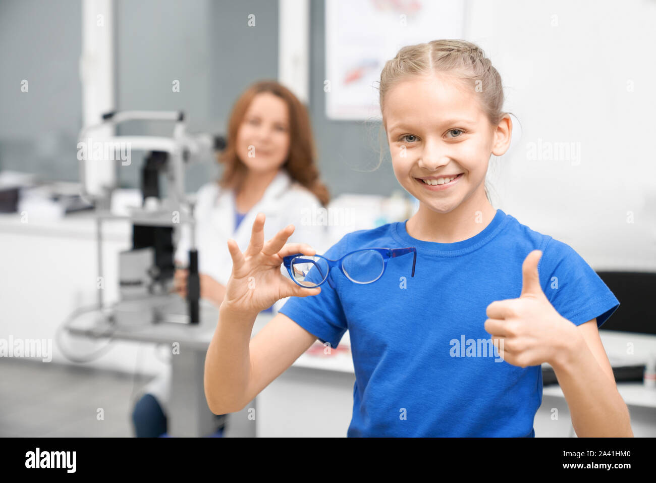 Vista frontale della bella ragazza in maglietta blu che mostra segno ok e acquisto di nuovi occhiali per visione. Client di giovani guardando la fotocamera e la scelta di occhiali in negozio di ottica. Concetto di oftalmologia. Foto Stock