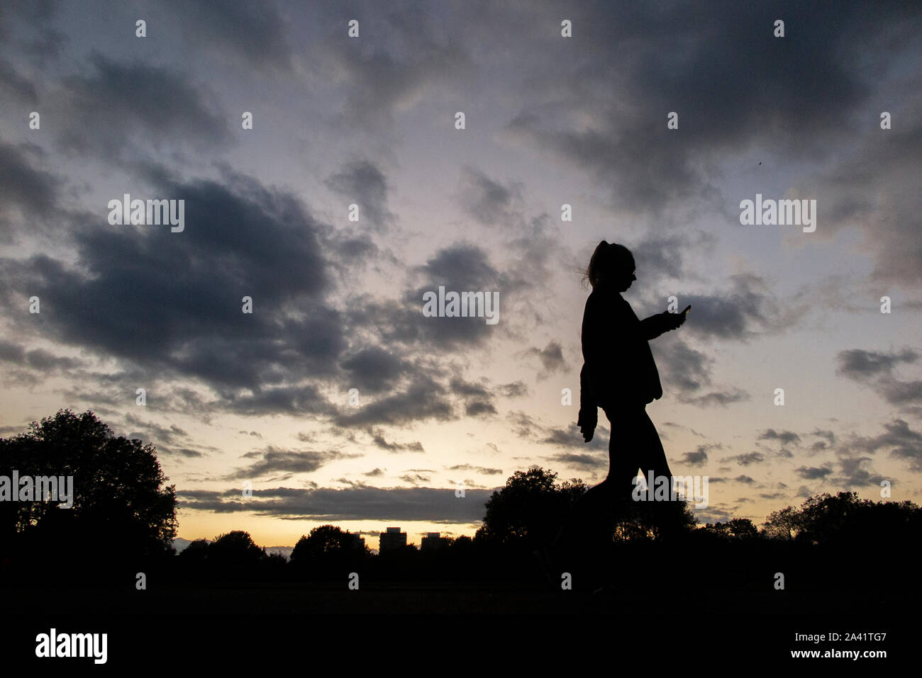Una singola ragazza cammina home, controllare il suo telefono cellulare in un tempestoso tramonto in un parco di Londra Foto Stock