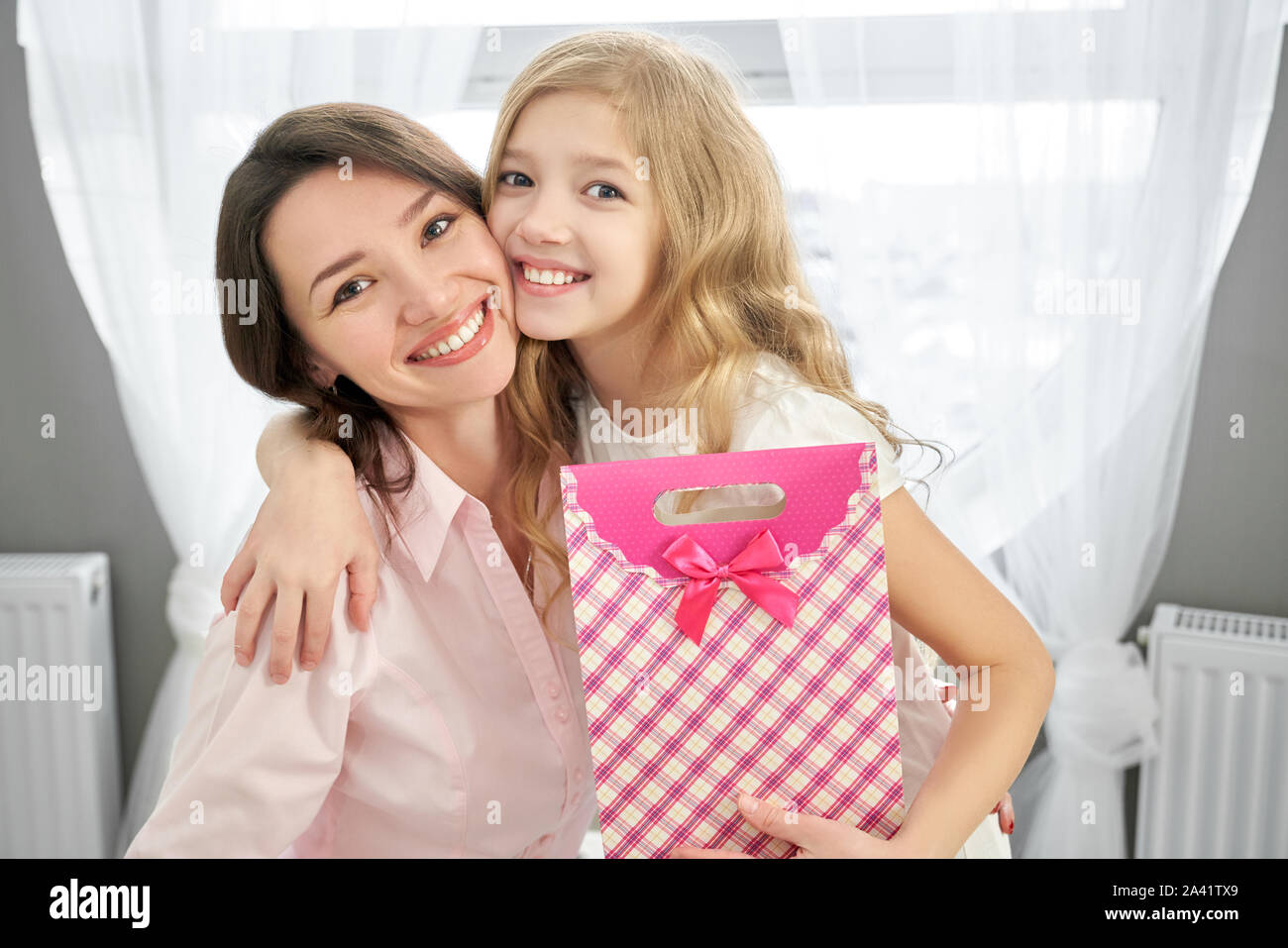Vista frontale di allegro madre e figlia piccola abbracciava, guardando la fotocamera e in posa con il presente. Cara ragazza presentando regalo per madre bella. Concetto di amore parentale e sorpresa. Foto Stock