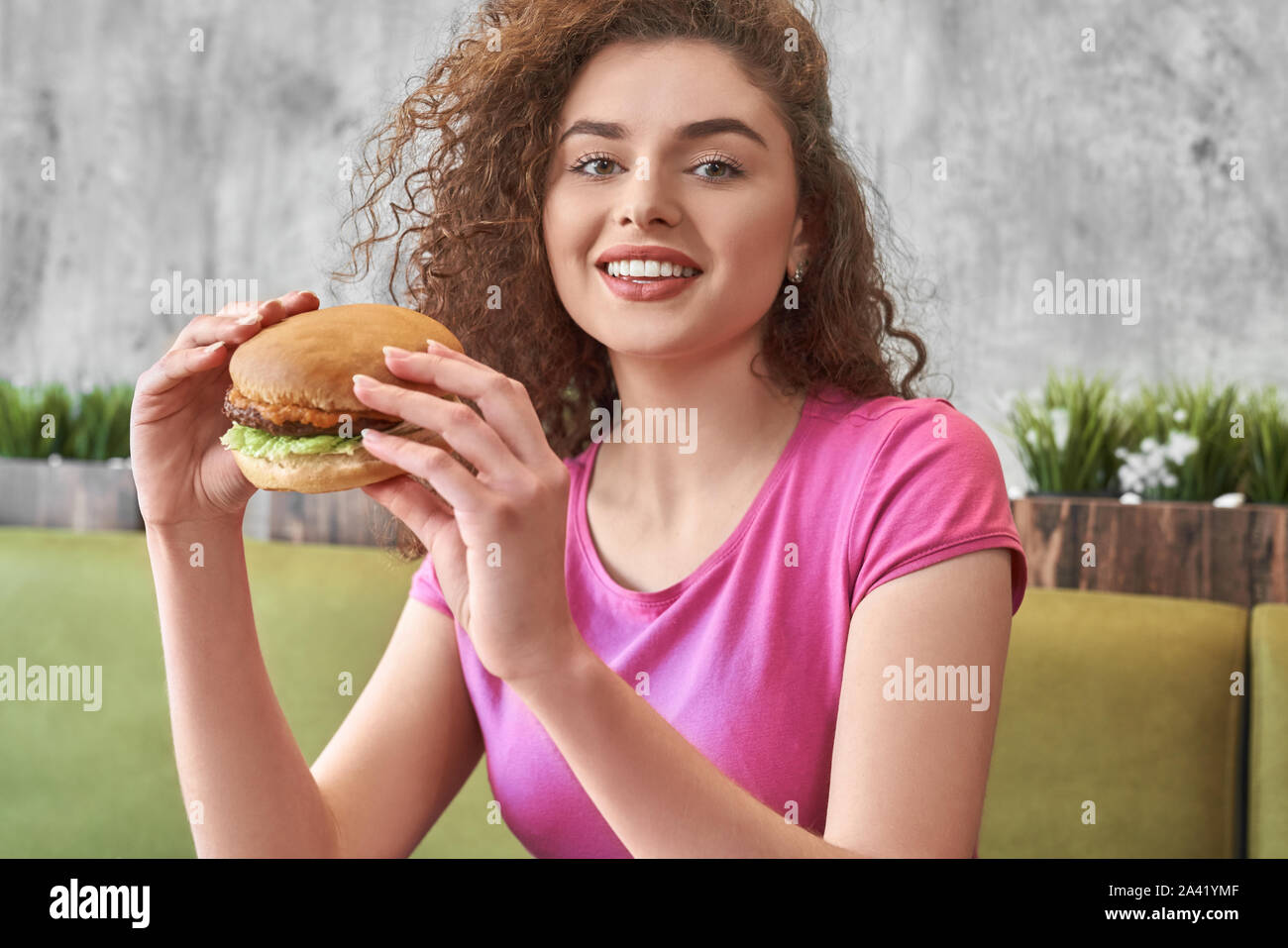 Vista frontale della fame bella donna che guarda la fotocamera mentre mangiare saporito grandi hamburger in cafe. Curly adolescente sorridente e in posa mentre godendo il fast food in pizzeria. Concetto di snack. Foto Stock