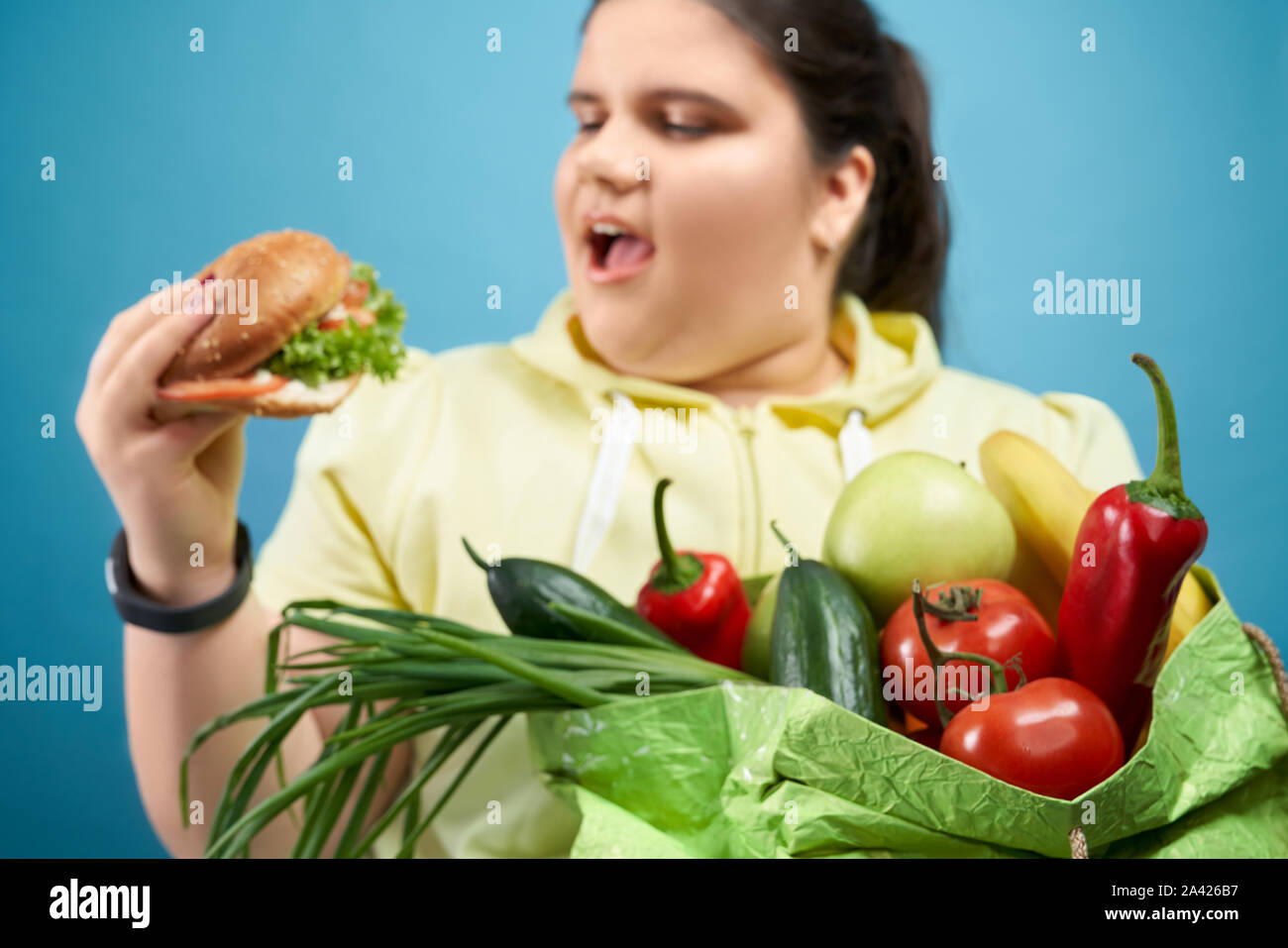 Lato del grasso brunette girl cercando di mangiare hamburger e mantenendo il pacchetto di frutta e verdura. Paffuto giovane femmina in felpa gialla è la scelta di un fast food invece di sani e freschi di pasto. Foto Stock