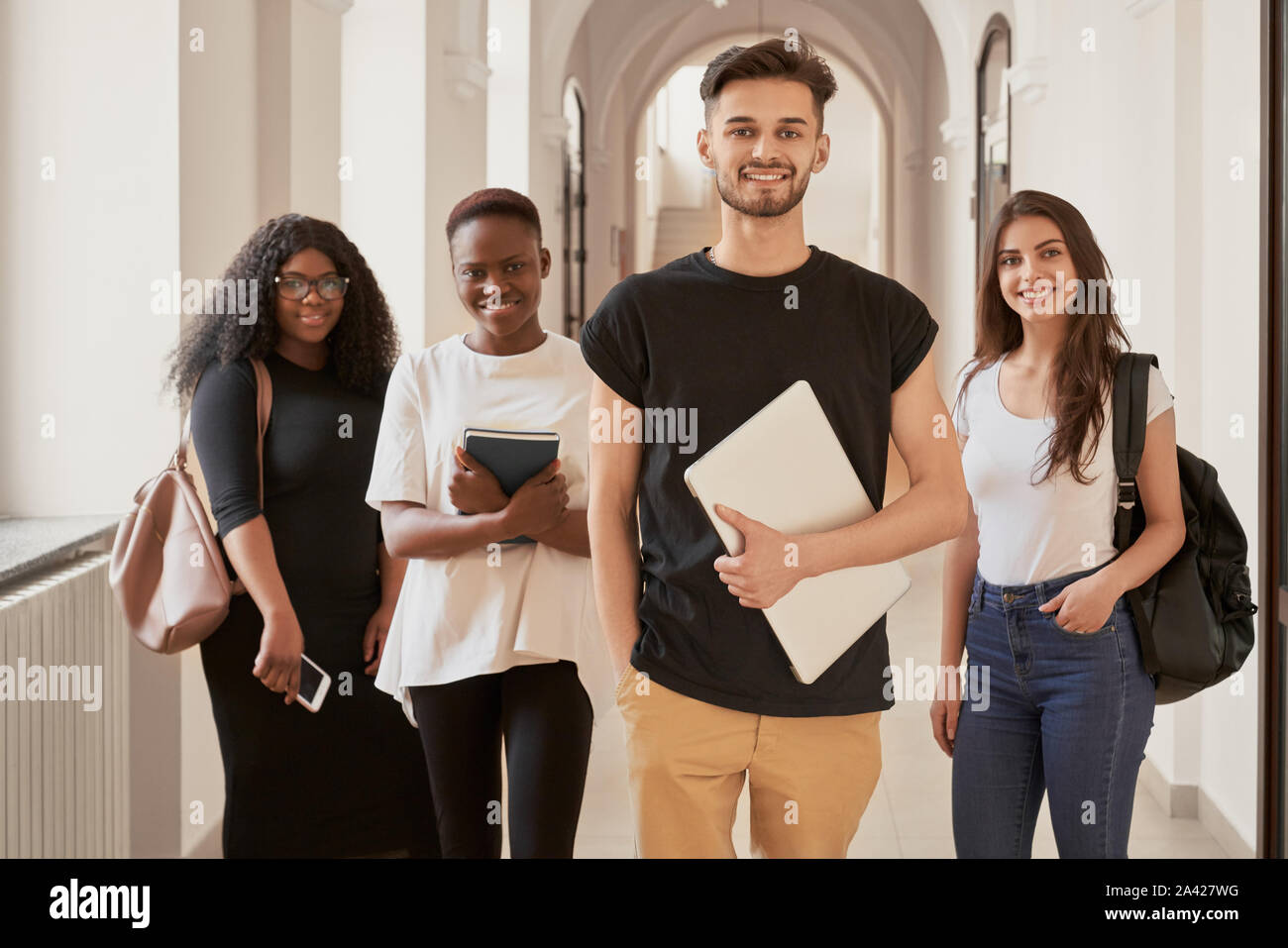 Multirazziale in gruppo di quattro studenti sorridenti vestiti casual in piedi nella sala universitaria,guardando la fotocamera e mantenere le loro cose personali.happy amici sta per rilassarsi dopo una dura giornata di studio Foto Stock