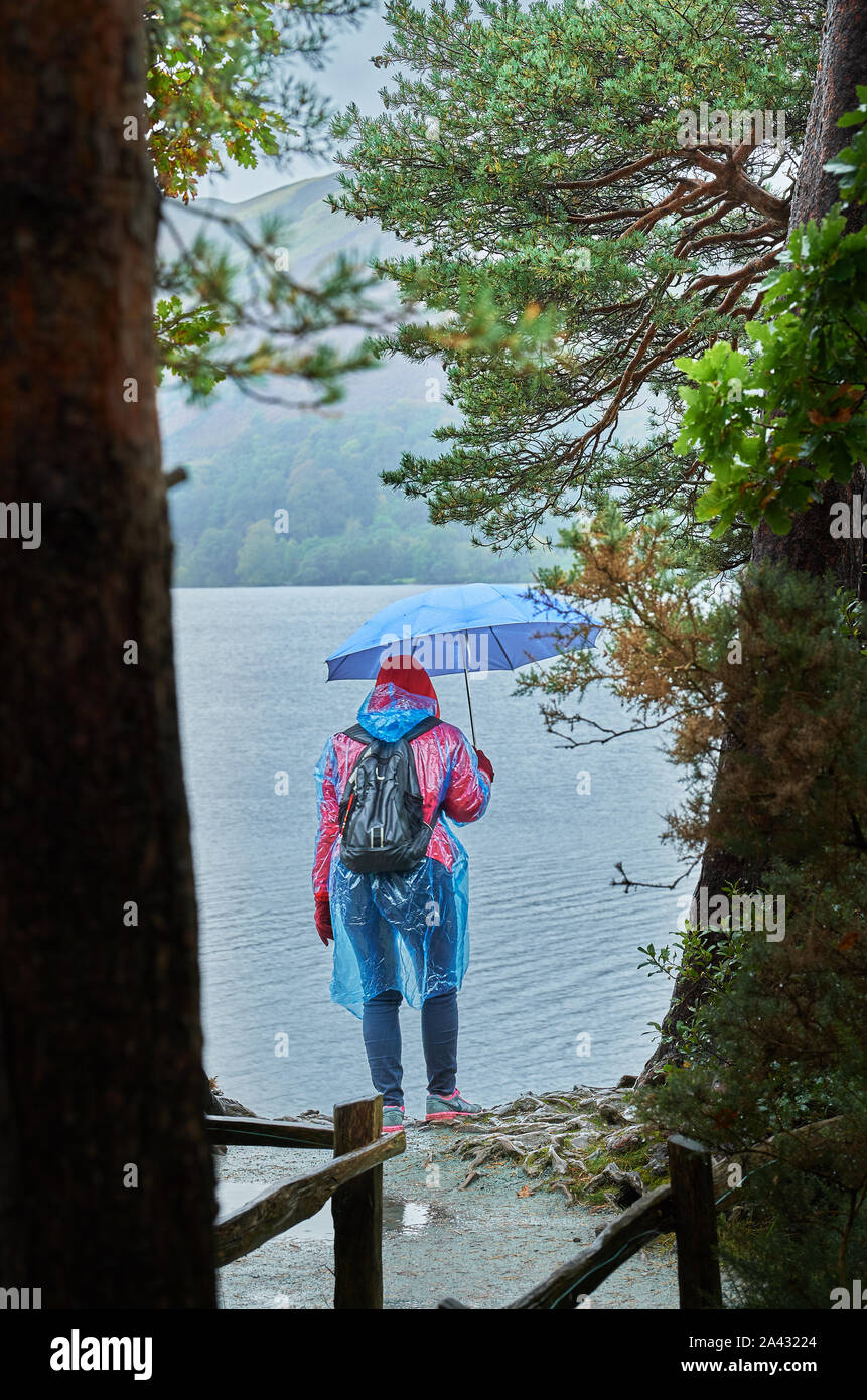 Una solitaria donna vestita di un mac seethrough e tenendo un ombrello sorge sul litorale su un nuvoloso, piovosa giornata al Lago Derwentwater, Lake District na Foto Stock