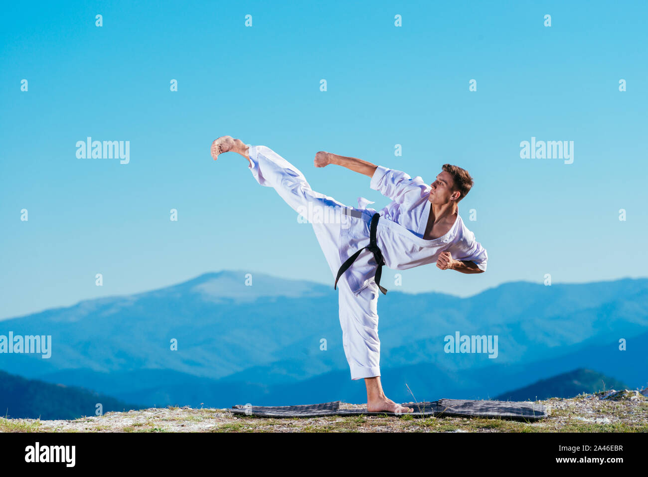 Il Karate uomo in un kimono esegue un lato gamba-piede kick(mae-geri) mentre si sta in piedi sul prato verde sulla cima di una montagna. Foto Stock