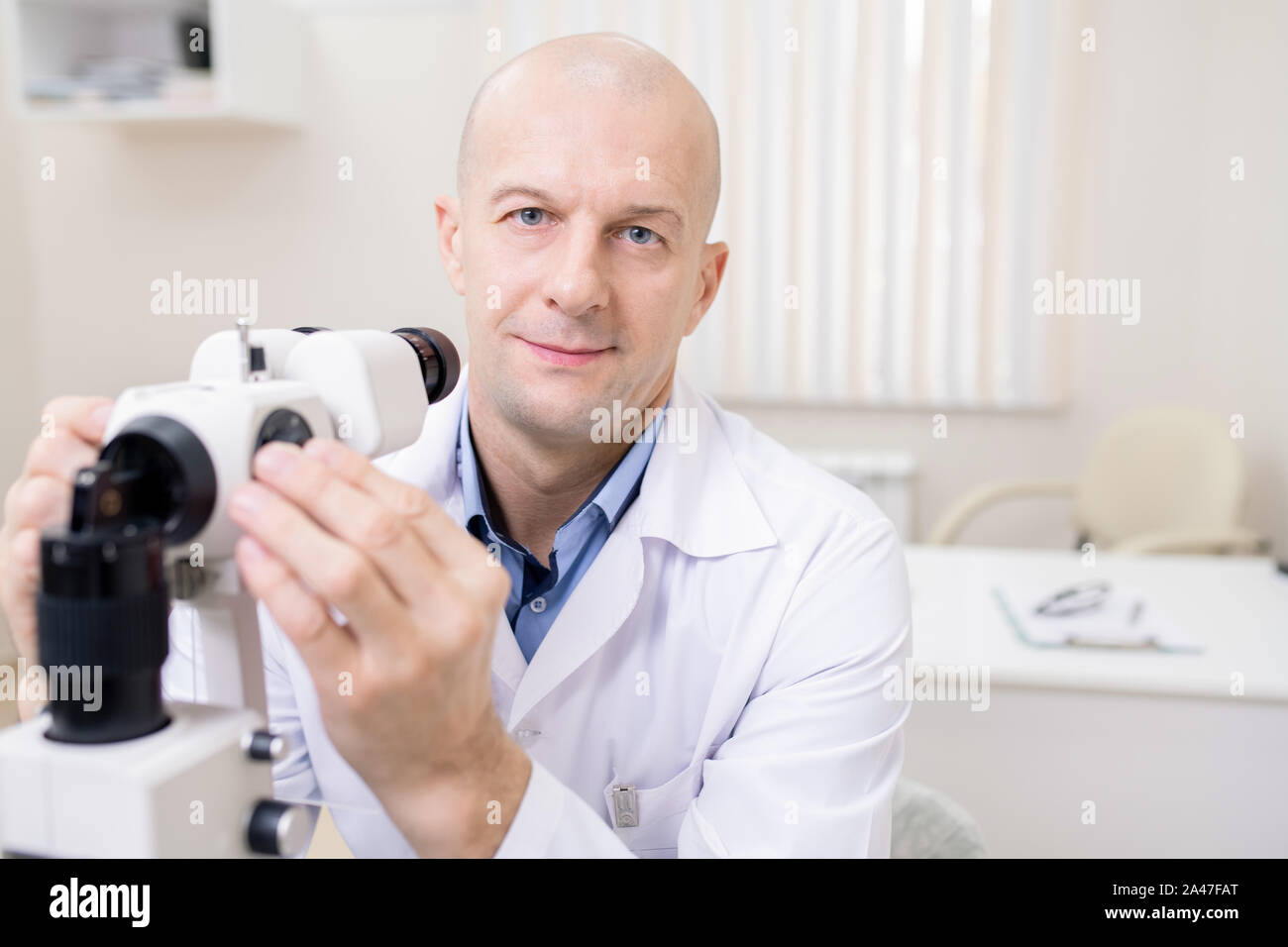 Bald di mezza età medico di occhio seduti nella parte anteriore della fotocamera dal luogo di lavoro Foto Stock