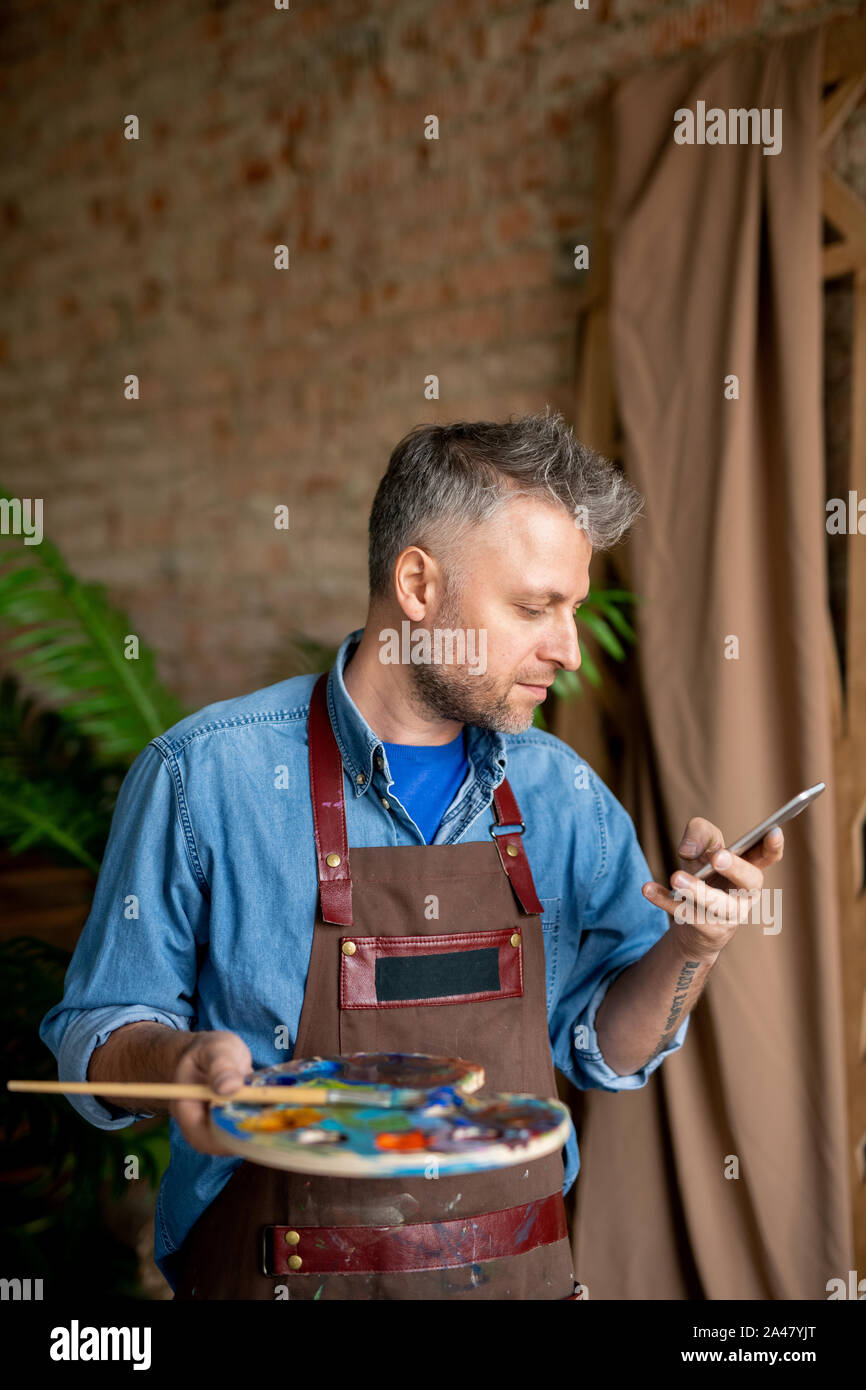 Artista serio in workwear guardando lo schermo dello smartphone mentre i messaggi di testo Foto Stock