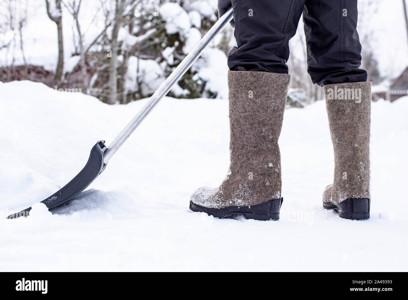 Il bidello in feltro scarpe con una pala si cancella una strada di campagna da un cumulo di neve, contro uno sfondo sfocato di una recinzione e alberi durante una connessione wi Foto Stock