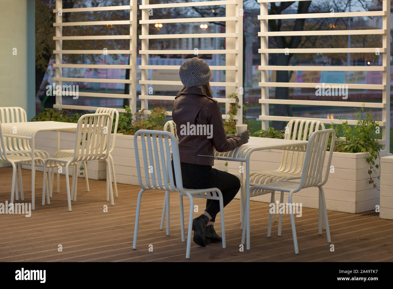 Un elegante ragazza è seduto lungo con caffè in street cafe. Una giovane donna sta godendo di caffè. In attesa di amici vista dal retro. Autunno piovoso evenin Foto Stock