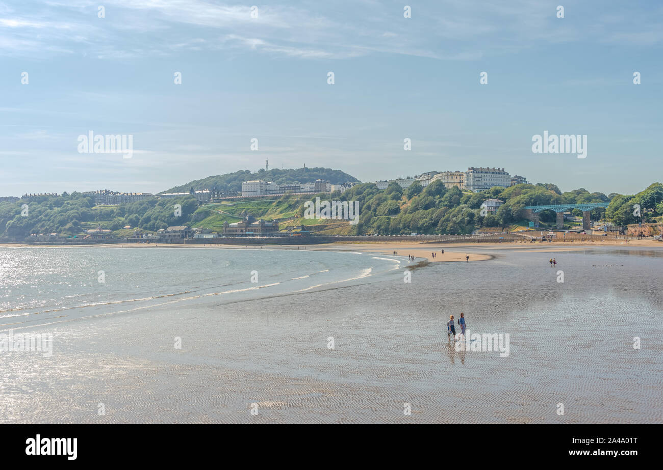 Persone godetevi il sole sulla spiaggia a Scarborough in tarda estate. Un hotel è accanto alla spiaggia come il promontorio si protende verso il mare. Un cielo blu è Foto Stock