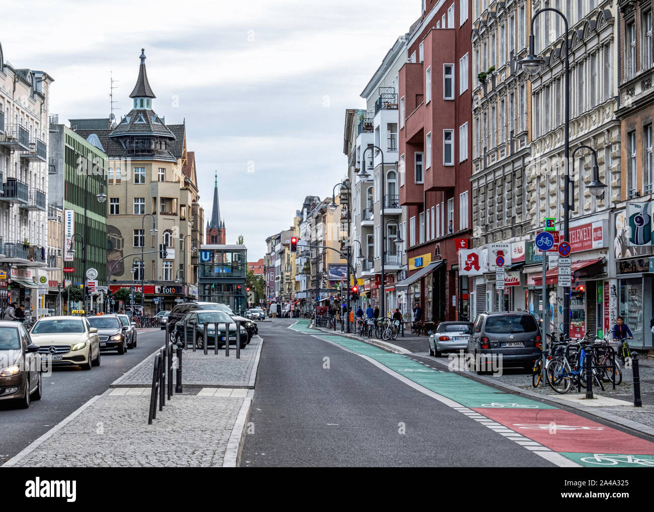 Vista sulla strada di negozi, palazzi, U-bahn ingresso & chiesa guglia in Karl Marx Strasse, Neukolln, Berlino Foto Stock