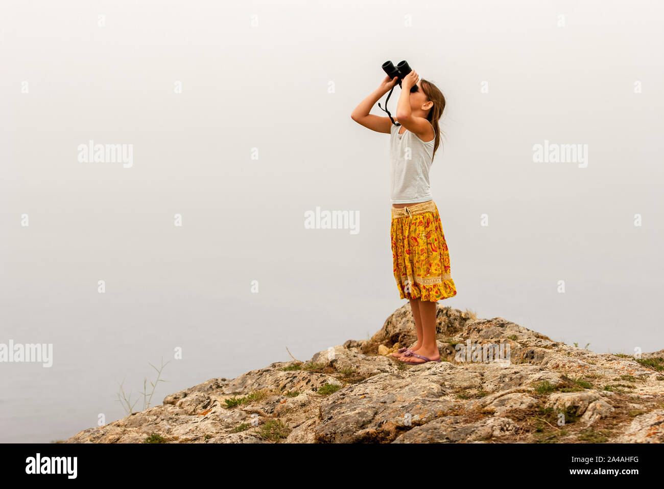 Bambino ragazza traveler sorge su una roccia sopra l'acqua e si guarda attraverso il binocolo in cielo. T-shirt e colorato di giallo gonna. Foto Stock