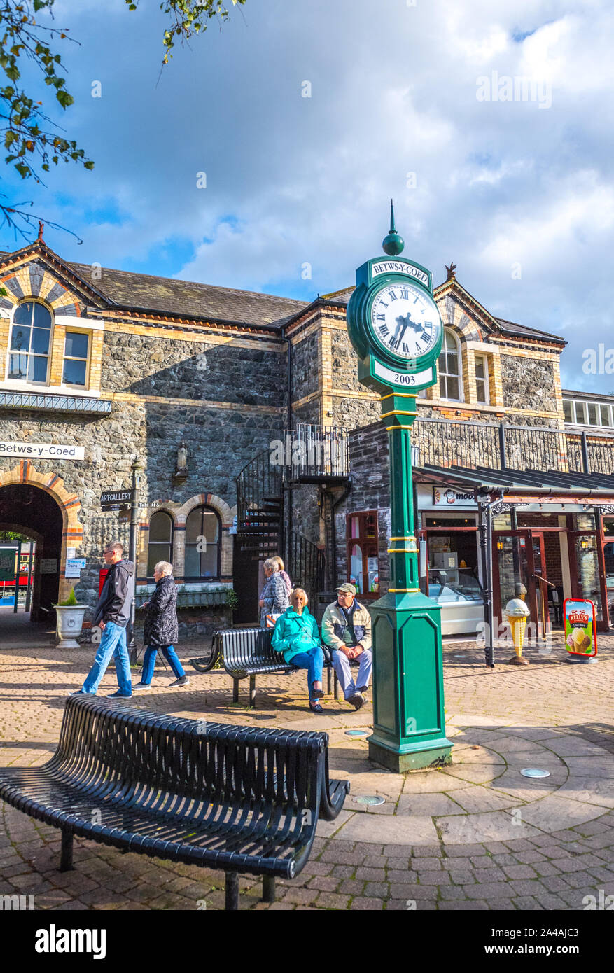 La gente seduta sulle panchine al di fuori del Betws-y-Coed stazione ferroviaria, Galles, con grandi ornati dipinte di verde orologio. Foto Stock