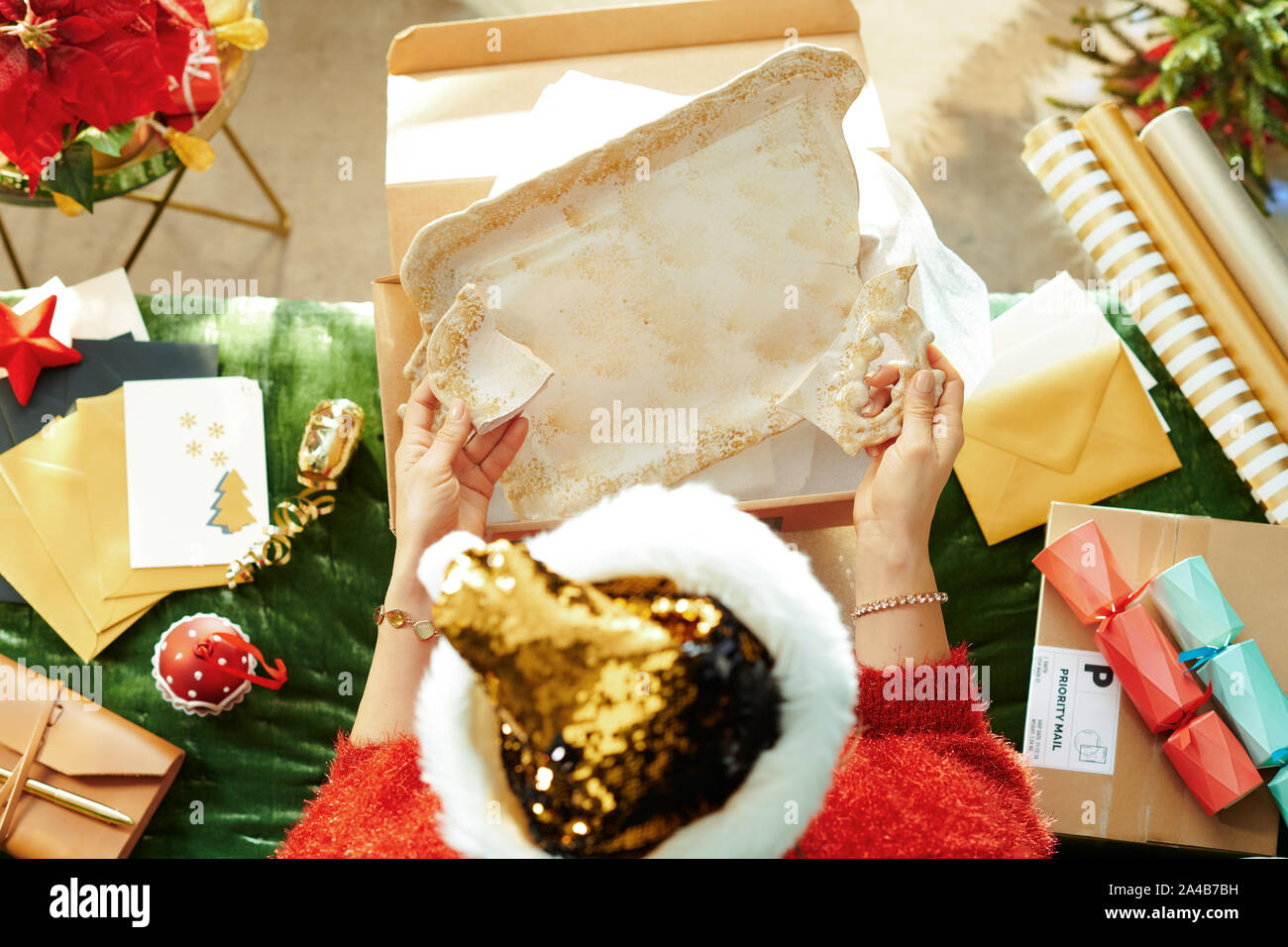 Stagione di vacanze. Vista superiore della donna moderna in sparkle oro zecchino santa hat ha ottenuto il pacco con piatto rotto, seduto sul divano del soggiorno moderno un Foto Stock