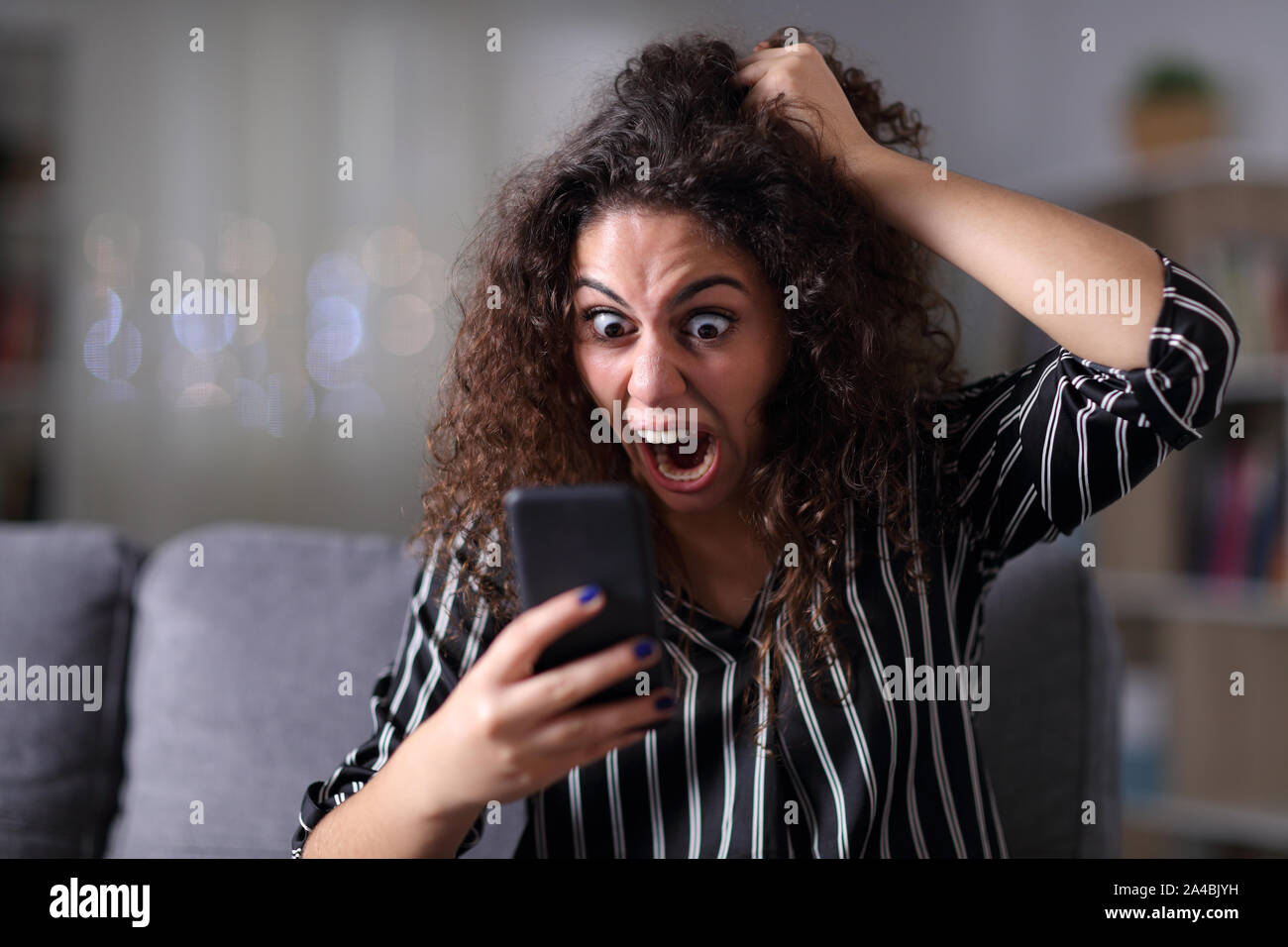 Donna isterica urlare il controllo smart phone content seduto su un divano nella notte a casa Foto Stock