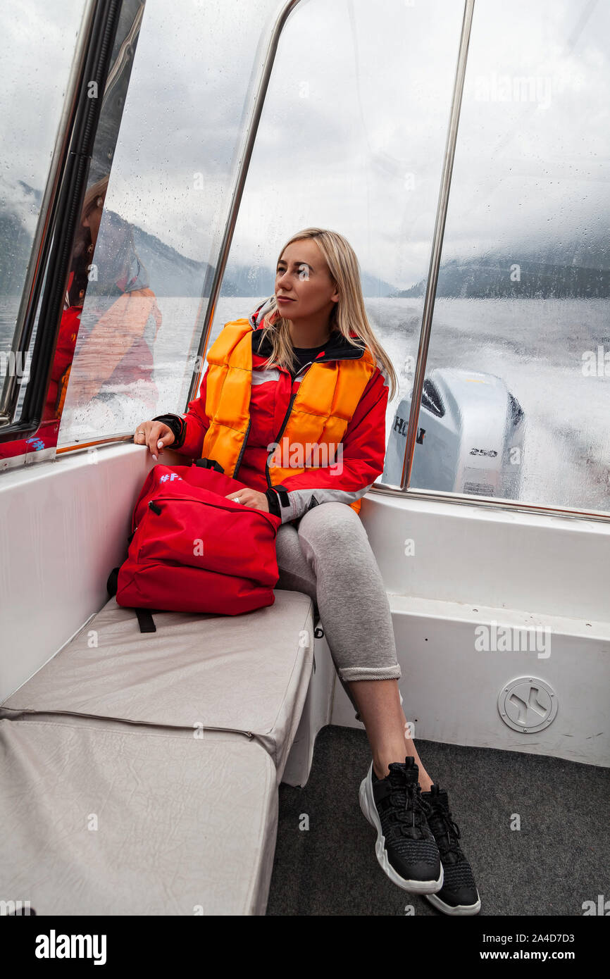 Giovane e bella ragazza bionda in un arancione giubbotto di salvataggio si siede in una barca a motore durante un escursione e la vela sul Lago Teletskoye nelle montagne di Altai. T