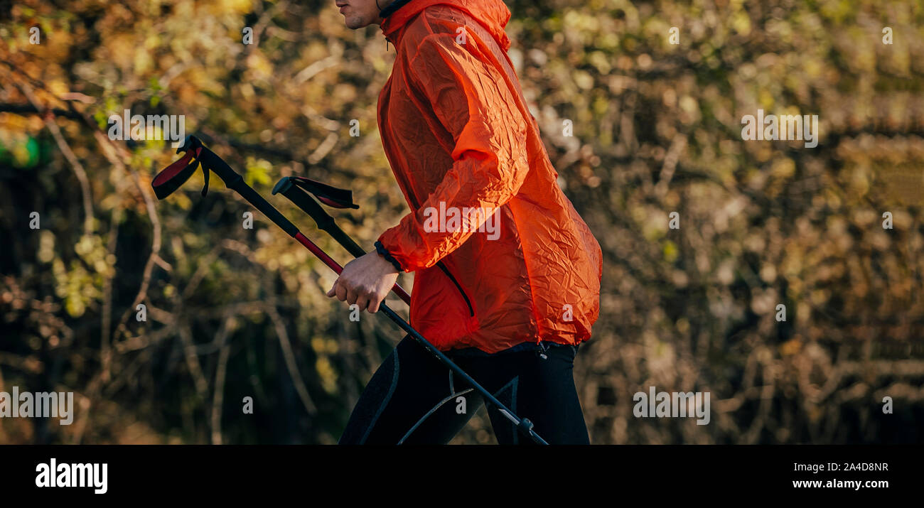 L'uomo runner con pali trekking in giacca rossa girare in background di autunno alberi con foglie di giallo Foto Stock