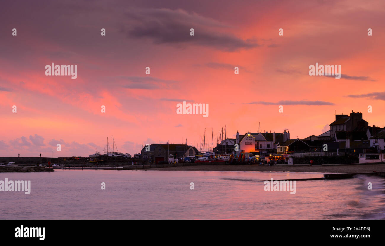 Lyme Regis, Dorset, Regno Unito. 13 ottobre 2019. Regno Unito Meteo: il cielo sopra il Cobb Harbout a Lyme Regis si illumina con gloriosi colori rosa e rosso al tramonto dopo un grigio e piovoso giorno. Credito: Celia McMahon/Alamy Live News. Foto Stock