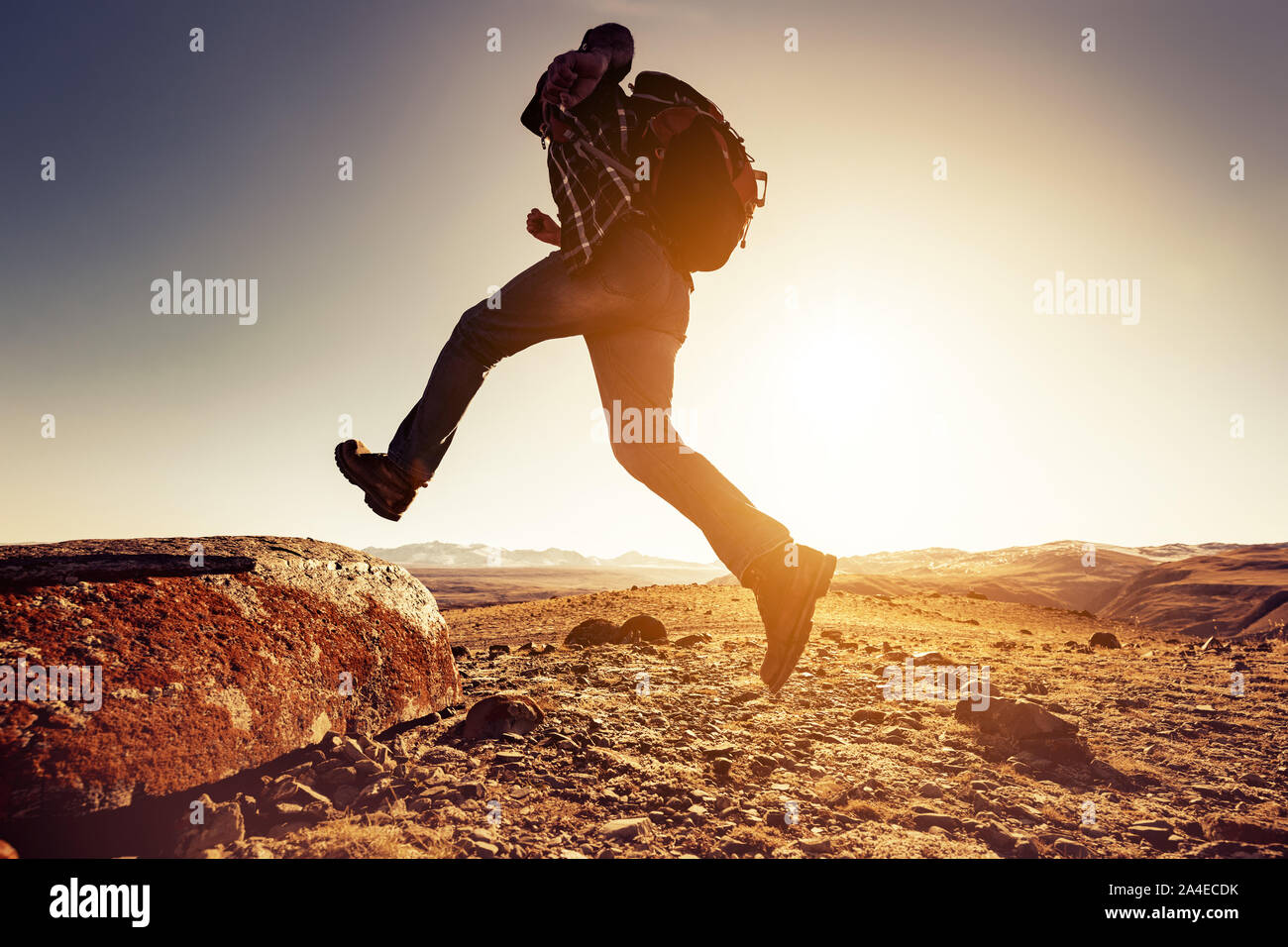 Escursionista uomo salta con grande zaino per un rock contro le montagne e il sole al tramonto Foto Stock