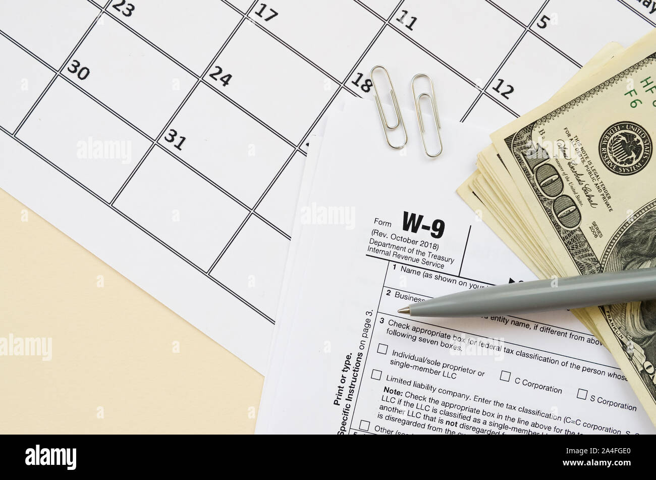 IRS Form W-9 richiesta per il contribuente un numero di identificazione e certificazione giace vuoto con penna e molte centinaia di fatture del dollaro nella pagina calendario. Imposta peri Foto Stock