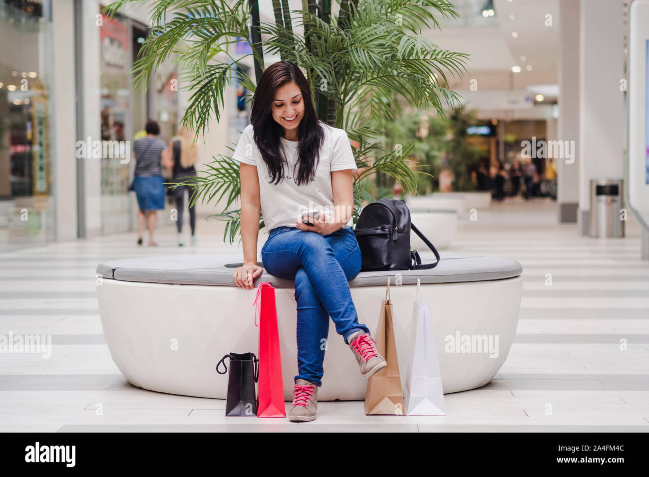 Donna seduta in mall guardando il suo telefono mobile e sorridente, con alcune borse della spesa sul pavimento. Il concetto di shopping. Foto Stock