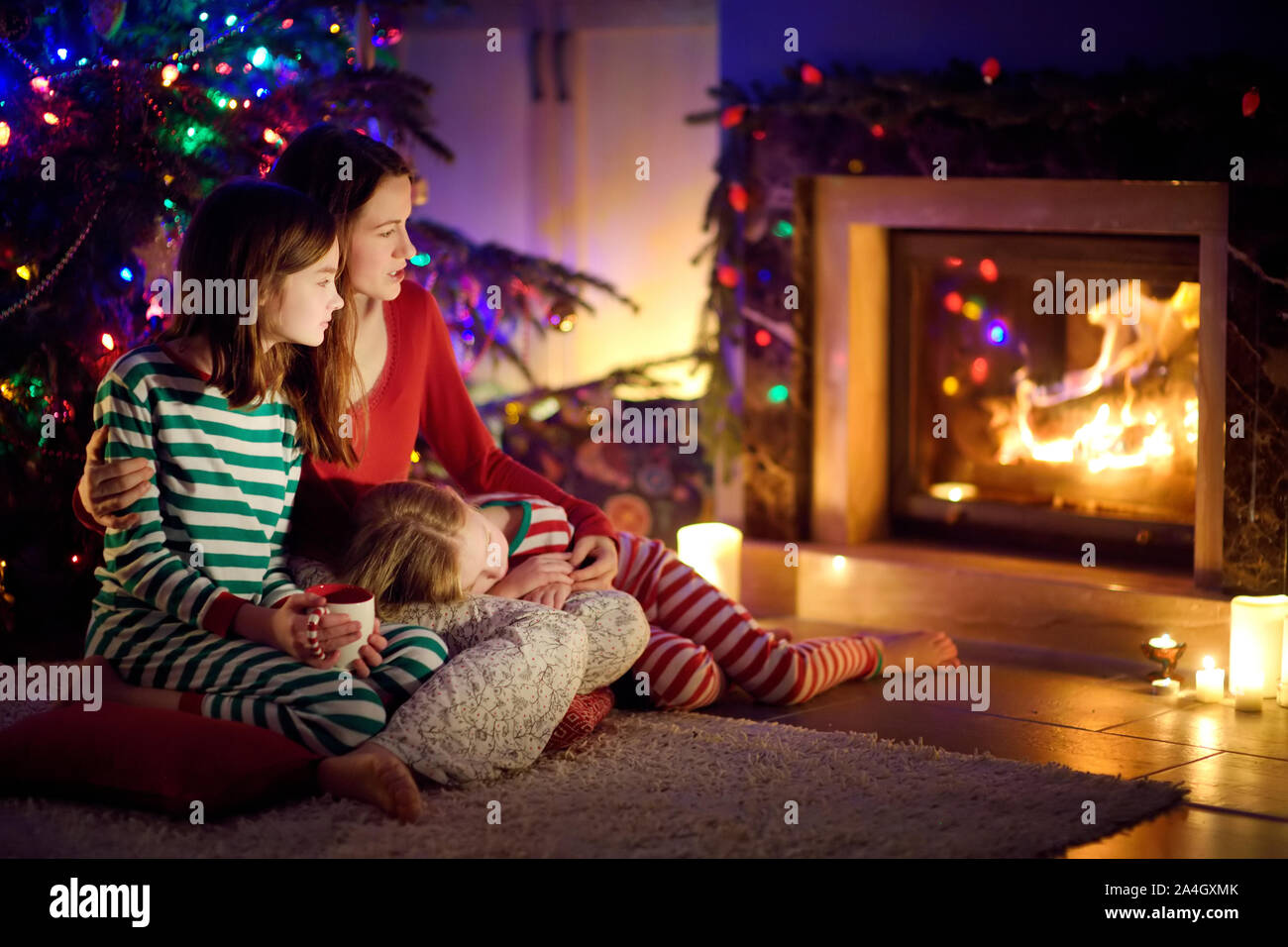 Felice giovane madre e le sue figlie avente un buon tempo seduti insieme da un camino in un buio accogliente soggiorno alla vigilia di Natale. Per celebrare il Natale Foto Stock