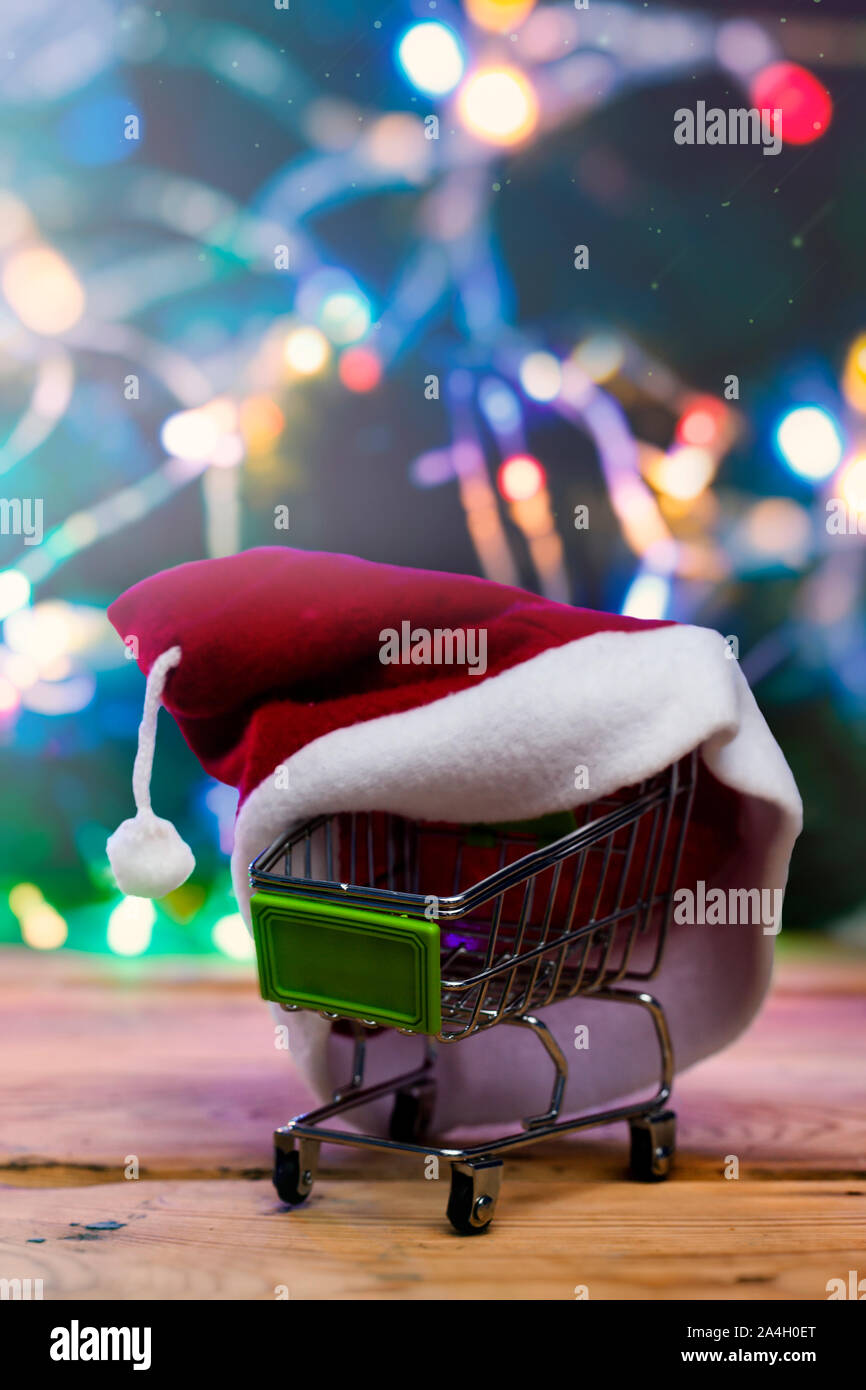 Shopping di Natale X-mas vendita concetto. Carrello spesa e Santa cap contro decorate con luci festose albero di Natale. Natale spendings. Inverno ho Foto Stock