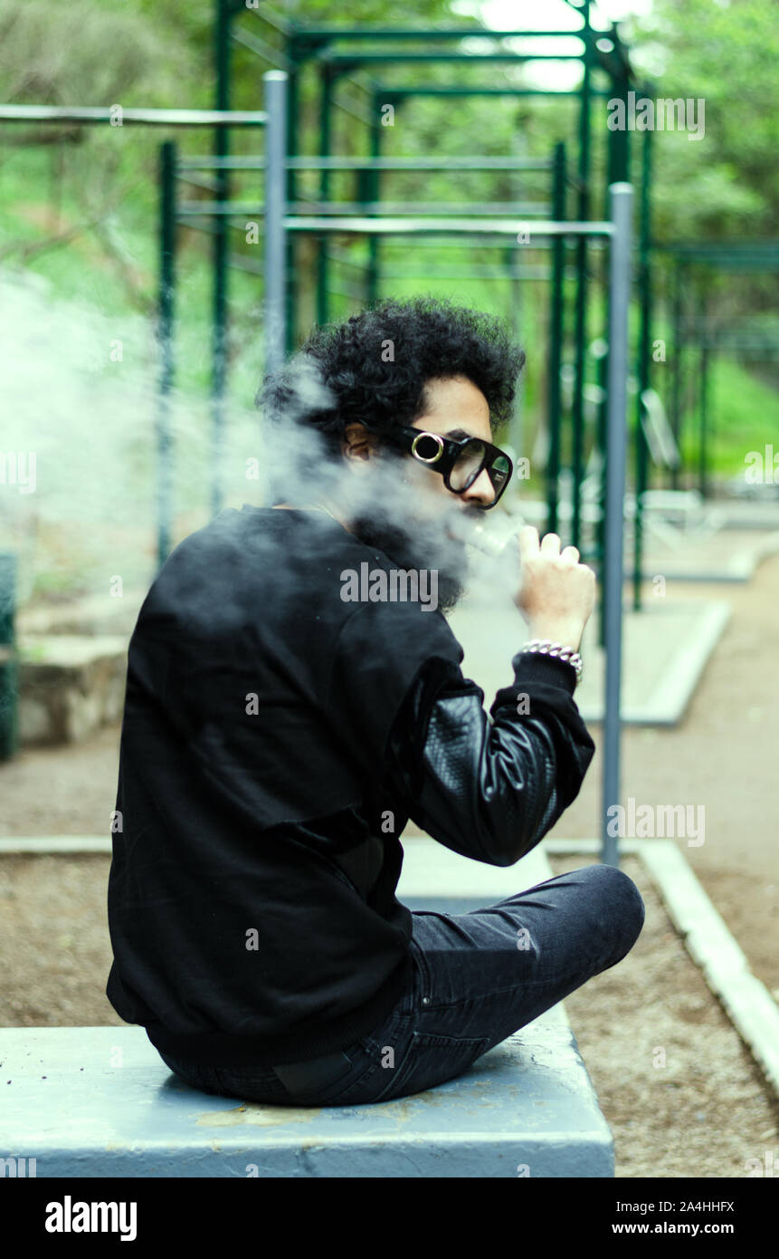 Con barba fuma una sigaretta elettronica a sedersi su una panchina nel parco Foto Stock