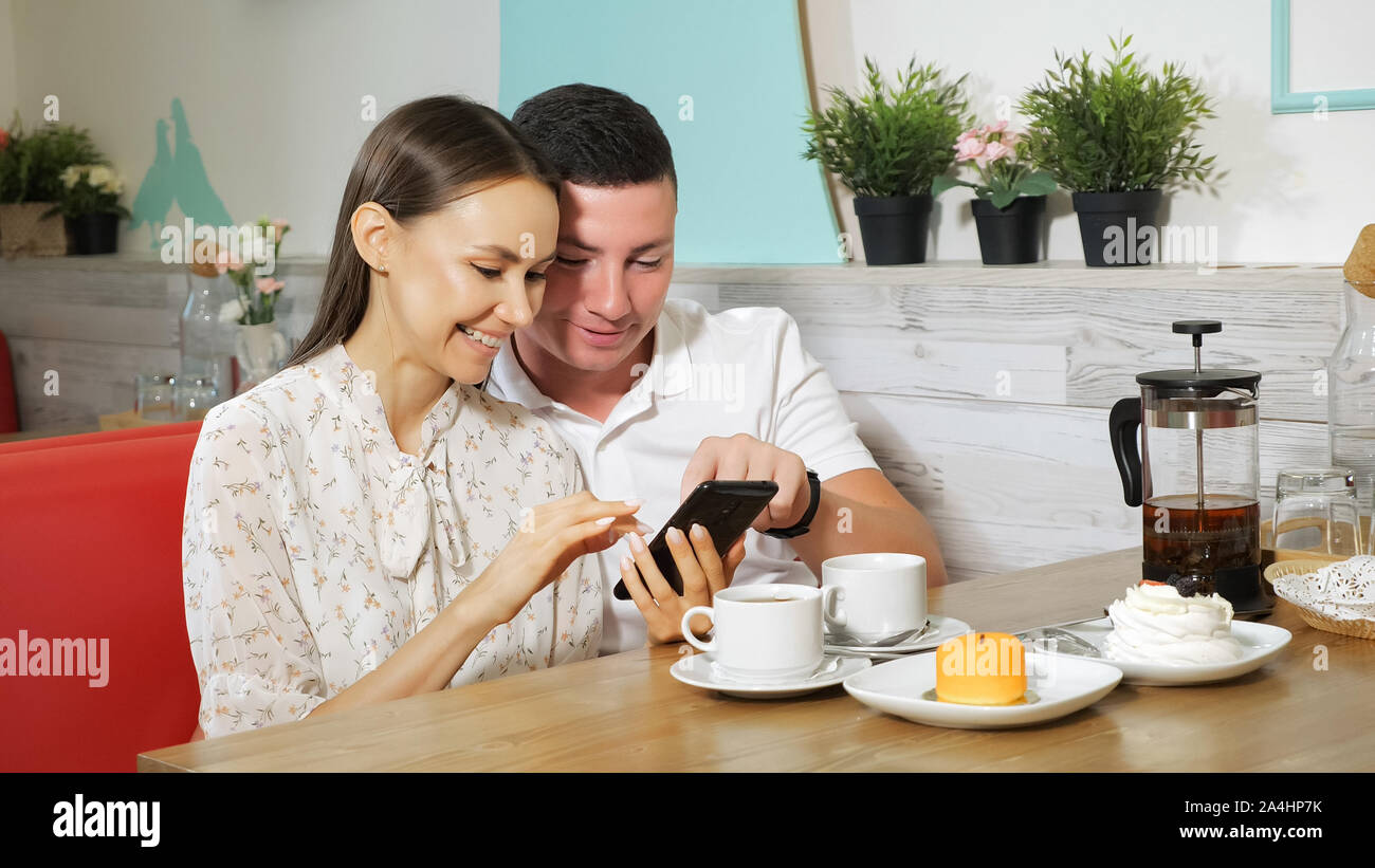 Ragazza gioiosa e guy guarda al moderno telefono cellulare al tavolo in legno con gustose torte e tè sulla tavola in accogliente negozio di dolci Foto Stock