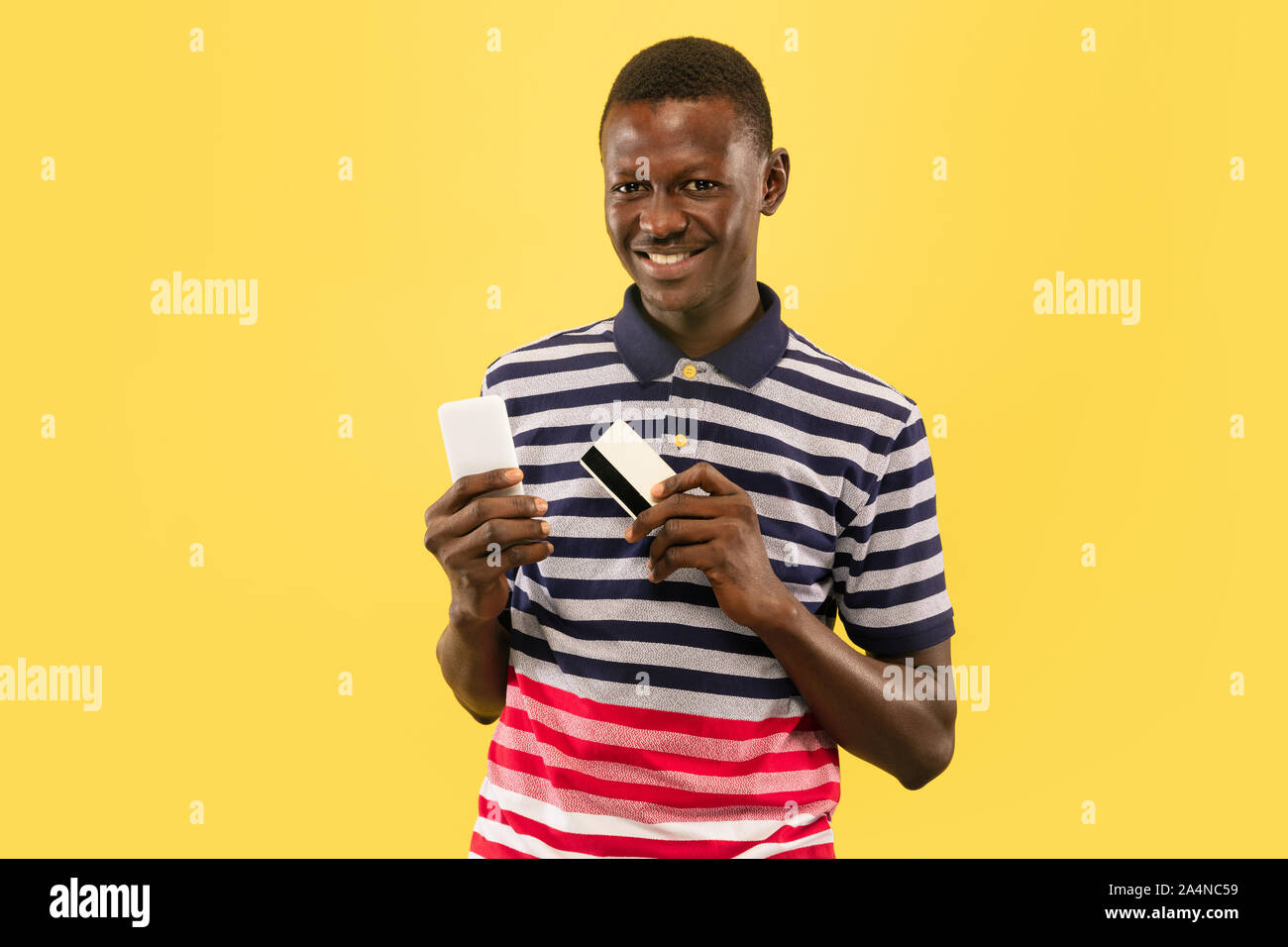 Giovani afro-americano con lo smartphone e scheda debet isolato giallo su sfondo per studio, l'espressione del viso. Bel maschio di mezza lunghezza verticale. Concetto di emozioni umane, l'espressione del viso. Foto Stock