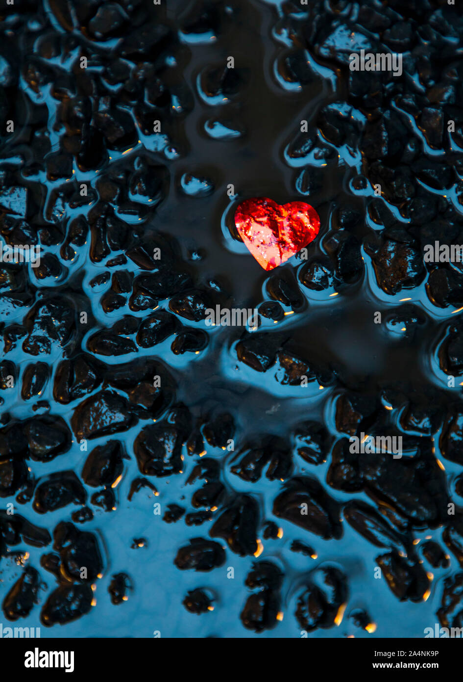 Un piccolo cuore rosso realizzato in stagnola, giace sul terreno bagnato, su pietre nere, i resti di un matrimonio, Foto Stock