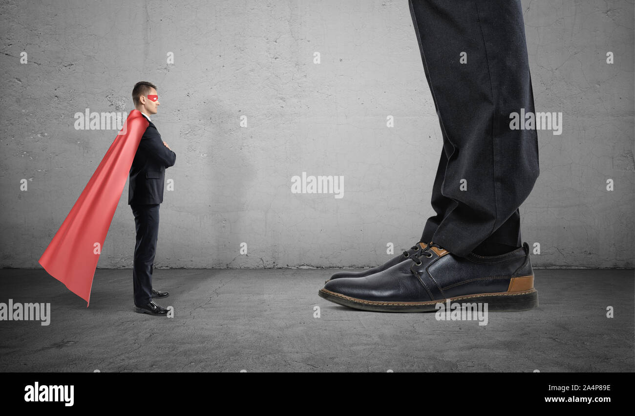 Un piccolo imprenditore in un supereroe cape sta di fronte l'uomo gigante con solo i suoi piedi visto. Foto Stock