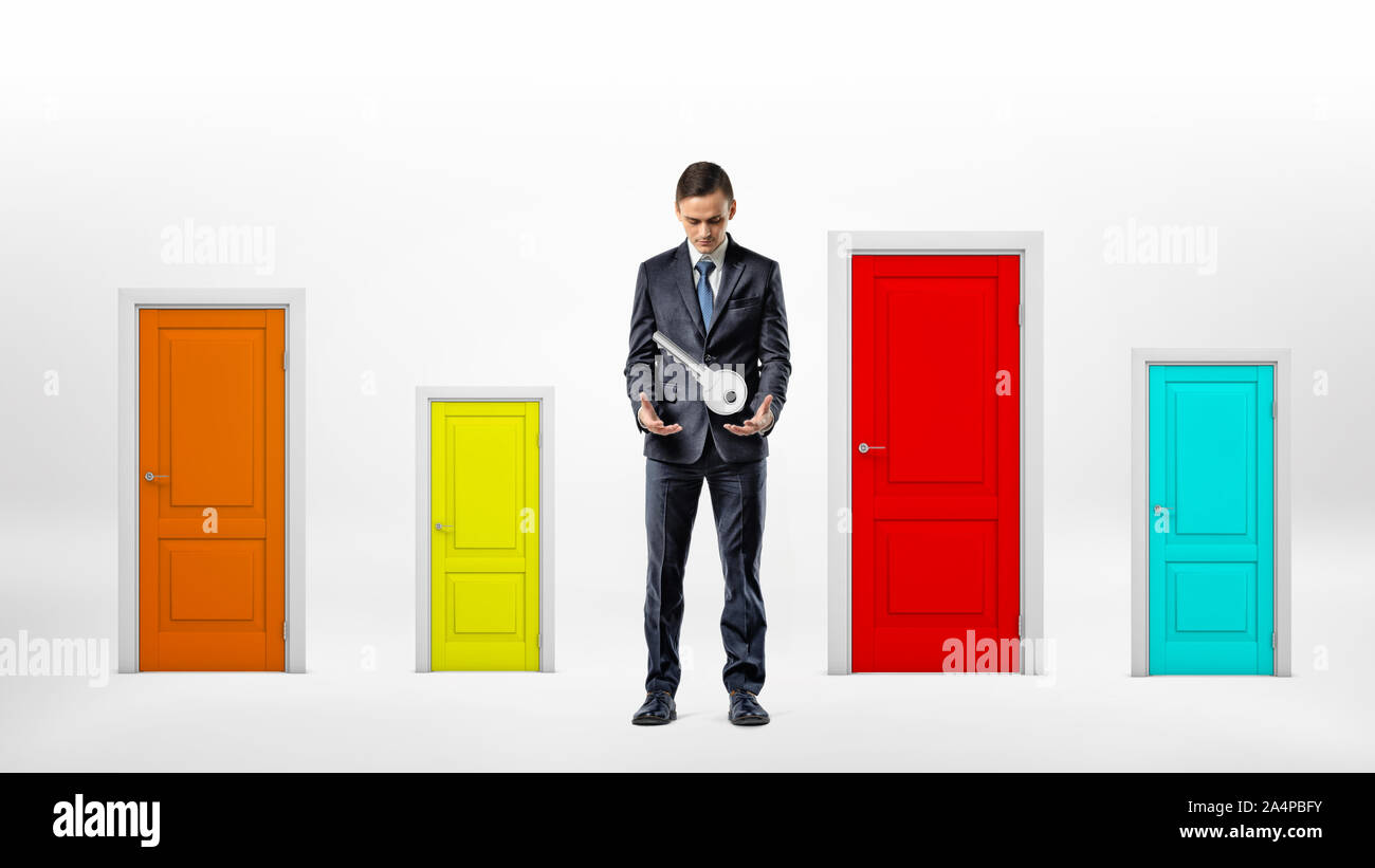 Un imprenditore si erge accanto a molte piccole porte multicolore e guarda a una chiave di grandi dimensioni appesi sopra le sue mani. Foto Stock