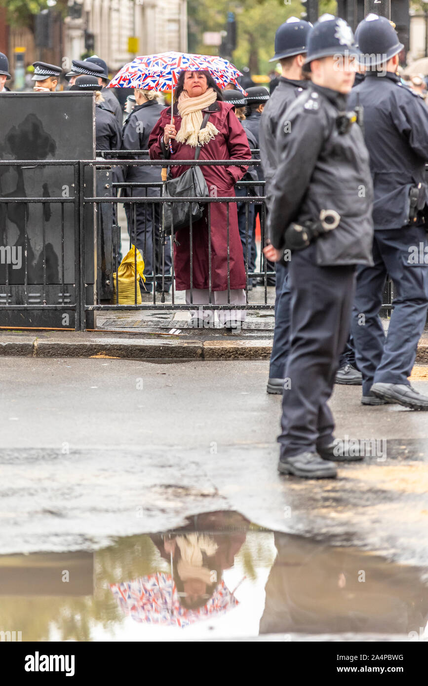 Lone spettatore femmina con britannica Union Jack flag ombrellone presso lo Stato apertura del Parlamento con un gran numero di forze di polizia. Wet Weather specchio di Pozza Foto Stock