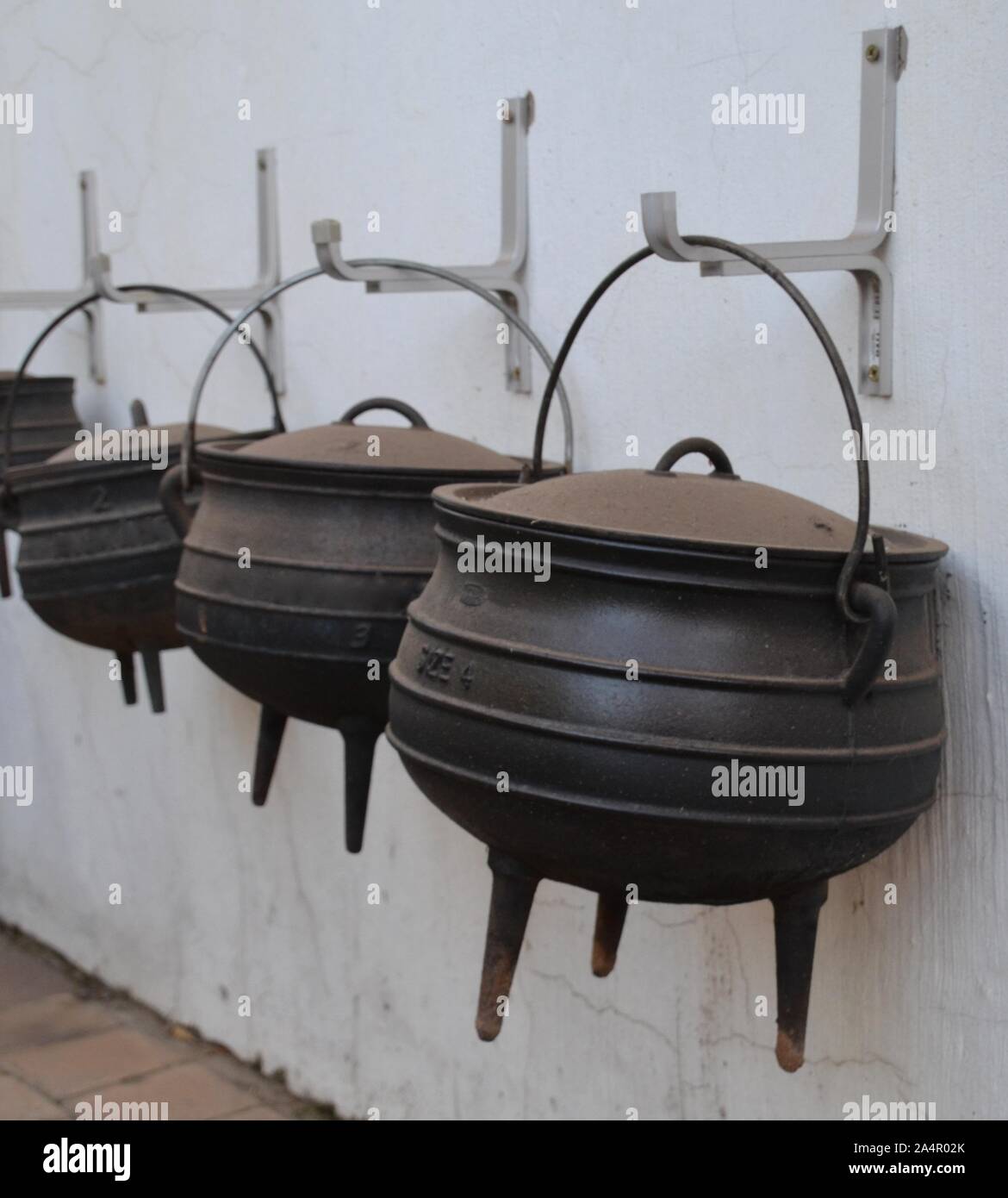 Linea di dimensione diversa ghisa tre zampe pentole di cottura appeso su un esterno muro dipinto di bianco in Sud Africa Foto Stock