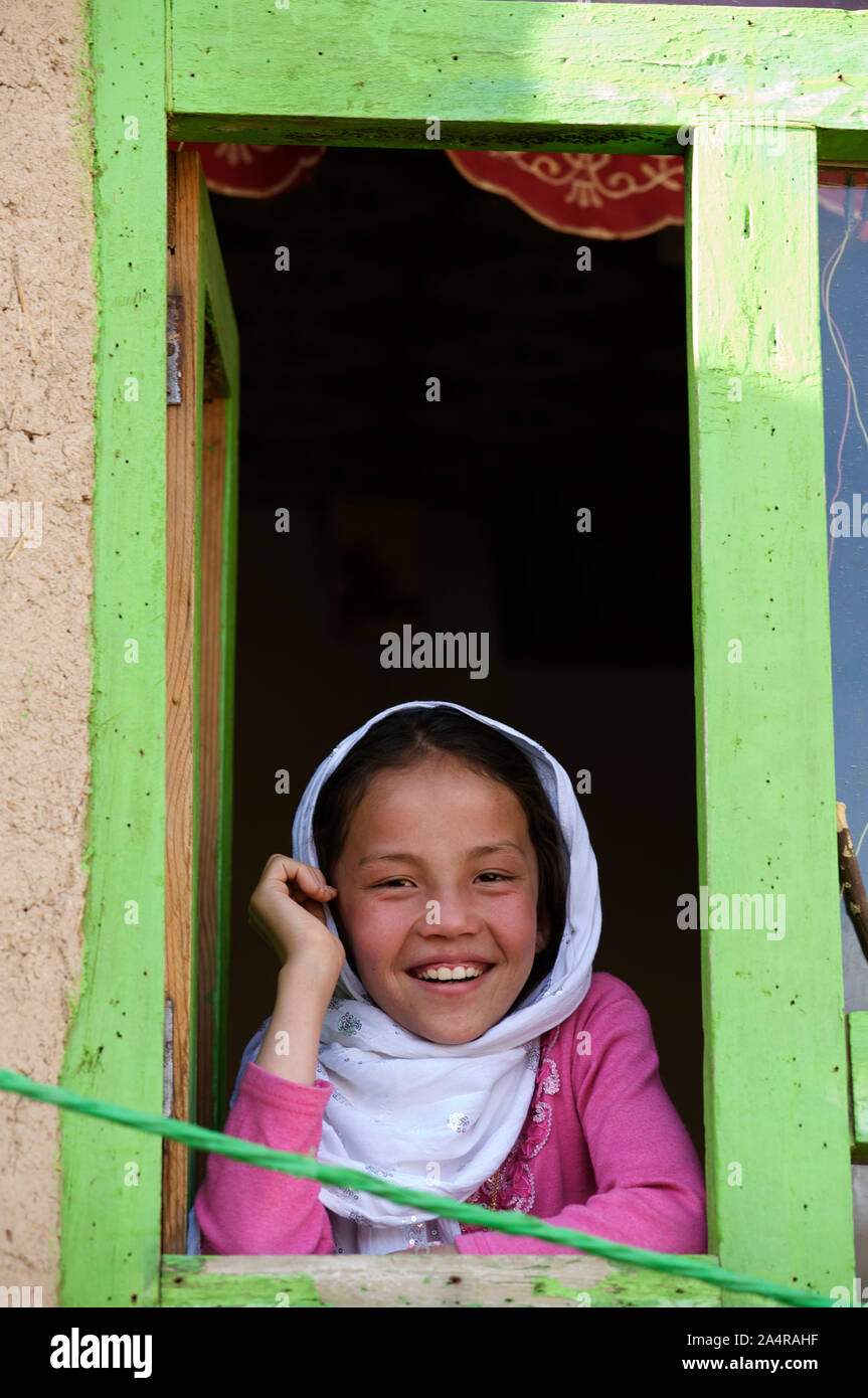 Un giovane afgano sorridente bambina guarda fuori dalla finestra di casa sua, nel villaggio di rag-e-Shad, nella periferia della città di Bamyan, centrale Provincia di Bamyan, Afghanistan. Il 11 maggio 2009. Foto Stock