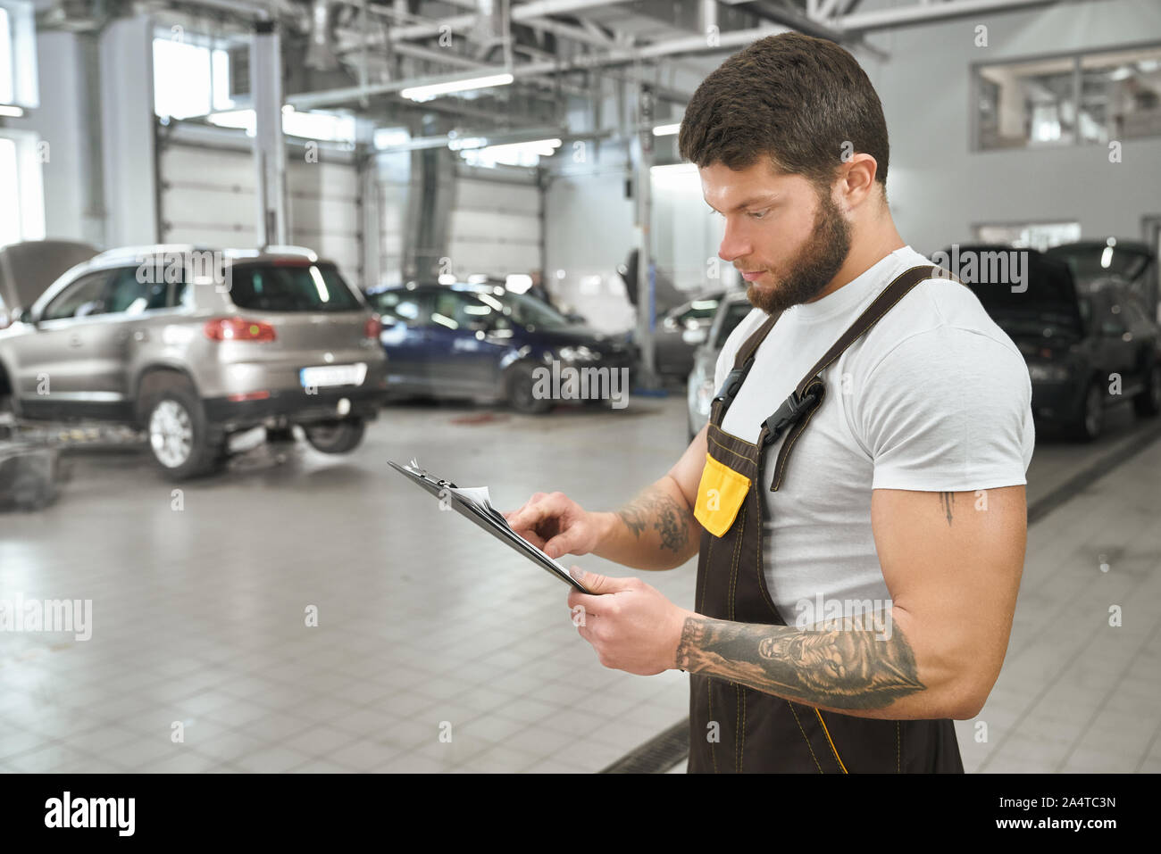 Bello, meccanica muscolare apposita cartella, Lettura. Uomo Barbuto con mani tattoed indossa in tute e white t shirt. Repairman lavorando in autoservice. Foto Stock