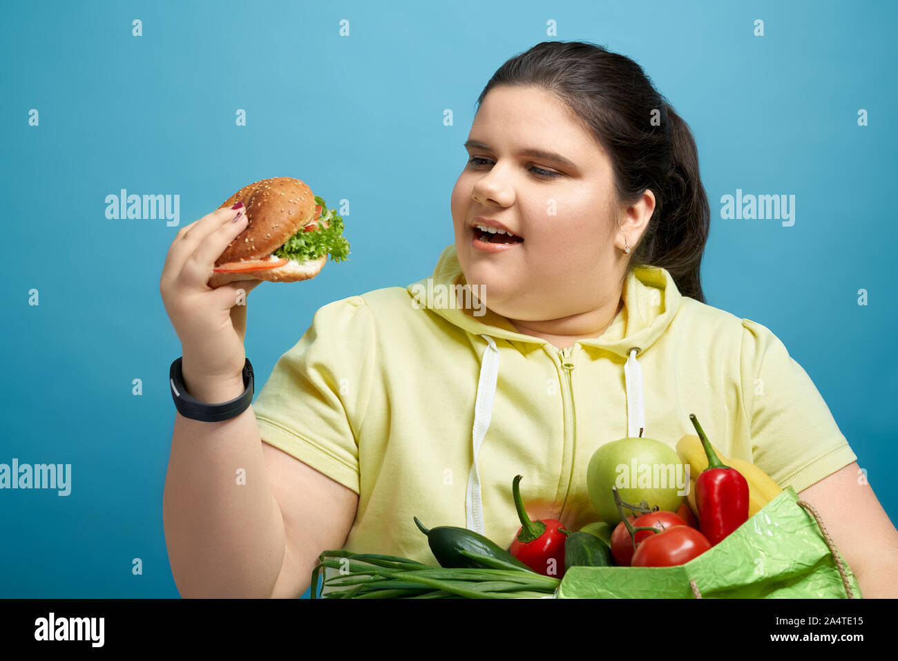 Chunky bruna giovane femmina è guardando hamburger con mouse aperto mantenendo la frutta e la verdura sulla sua mano. Concetto di temporale tentazione malsana invece di scegliere il mangiare sano. Foto Stock