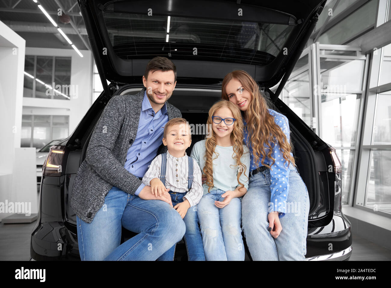 Vista frontale dei dolci famiglia seduti insieme sul tronco su auto, guardando la telecamera, avvolgente e in posa di auto salon. Genitori felici con i bambini la scelta e acquisto di nuove automobili confortevoli. Foto Stock