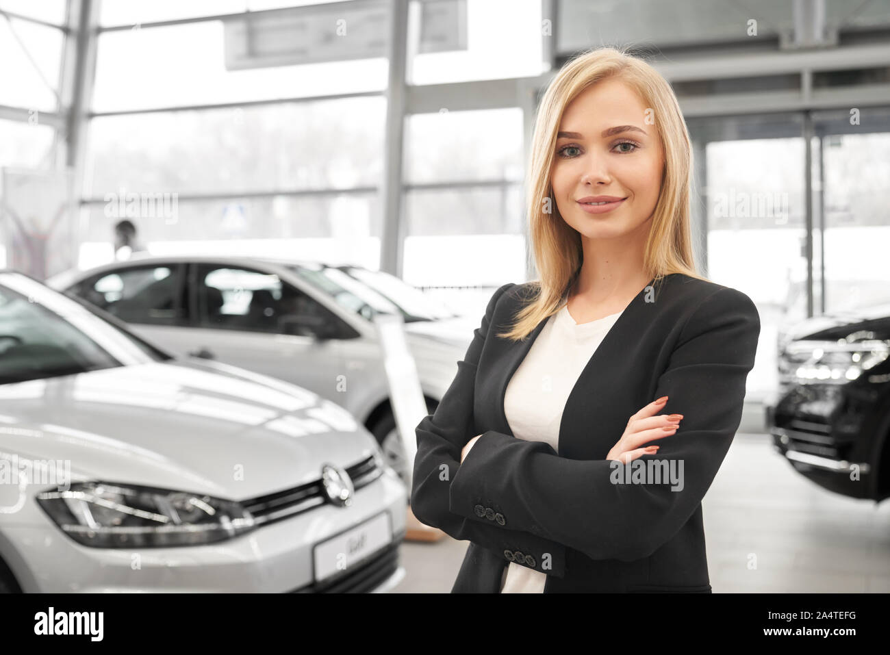 Vista frontale della femmina attraente rivenditore di auto con bracci ripiegati in piedi in auto salon, guardando la fotocamera e in posa. Bravo lavoratore la vendita di automobili nuove e ad aiutare i clienti in showroom. Foto Stock