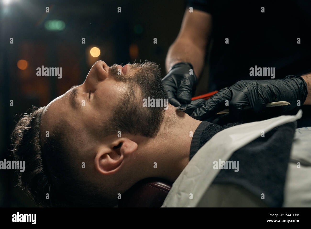 Vista laterale del barbuto bello ragazzo sdraiato sul divano mentre competenti barbiere maschio in guanti neri la rasatura la sua barba da rasoio affilato.client di giovani al barbiere è cambiare acconciatura e aspetto Foto Stock