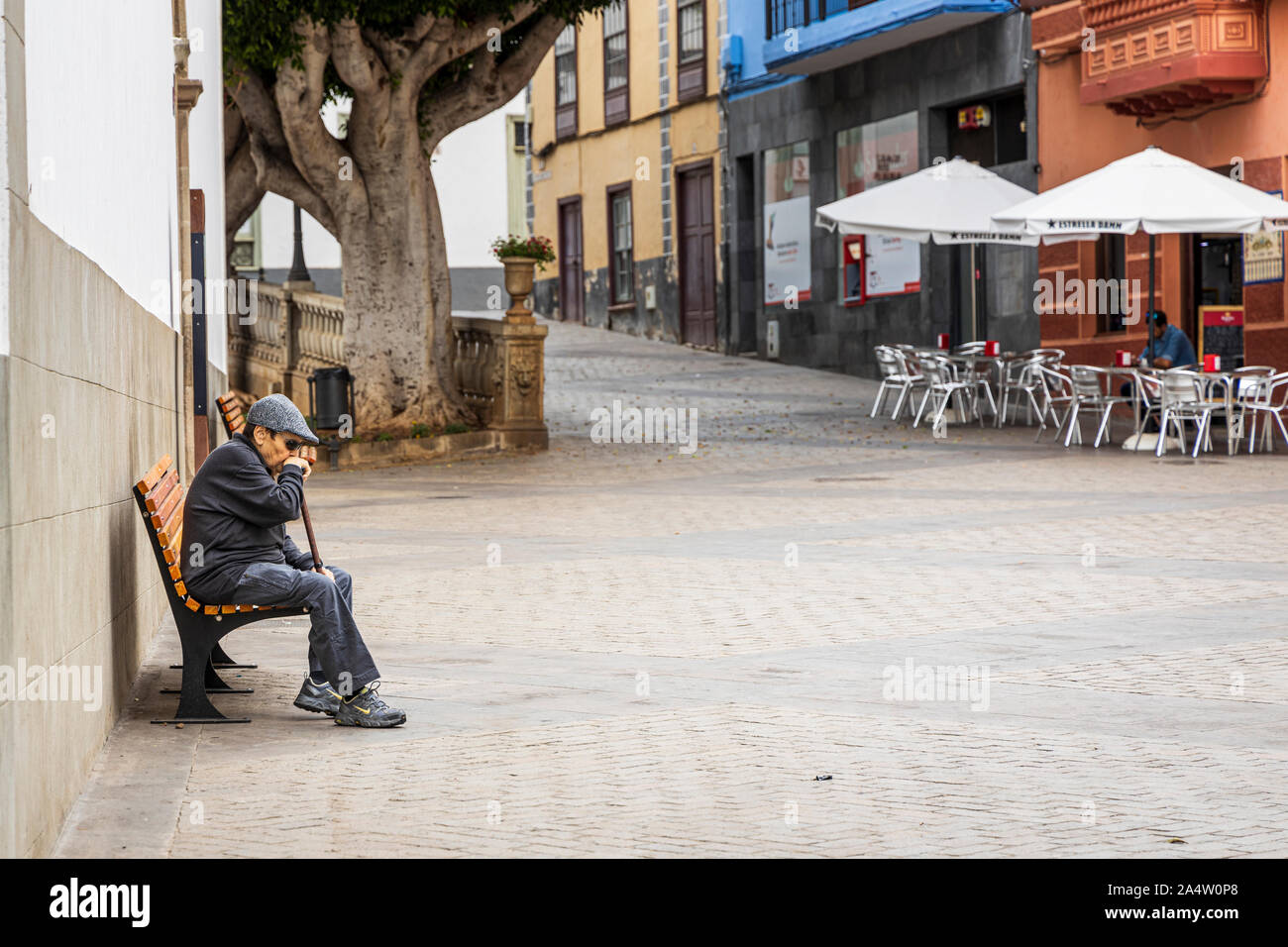 Vecchia persona con tappo piatto, occhiali scuri seduta su una panchina pensively pendente su un bastone da passeggio in Plaza a Guia de Isora, Tenerife, Isole Canarie Foto Stock