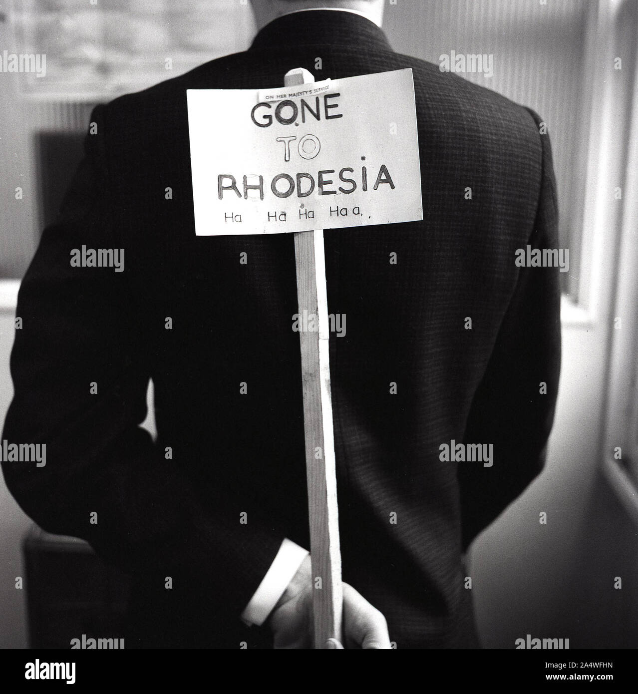 Anni '1960, storico, all'interno di un ufficio, un uomo che tiene un bastone di legno dietro la schiena, con un biglietto su di esso che dice "andato in Rhodesia", ha ha ha ha...... La Rhodesia fu uno stato non riconosciuto nell'Africa meridionale tra il 1965 e il 1979, dopo aver dichiarato una dichiarazione di indipendenza unilaterale (UDI) nel novembre 1965. Foto Stock