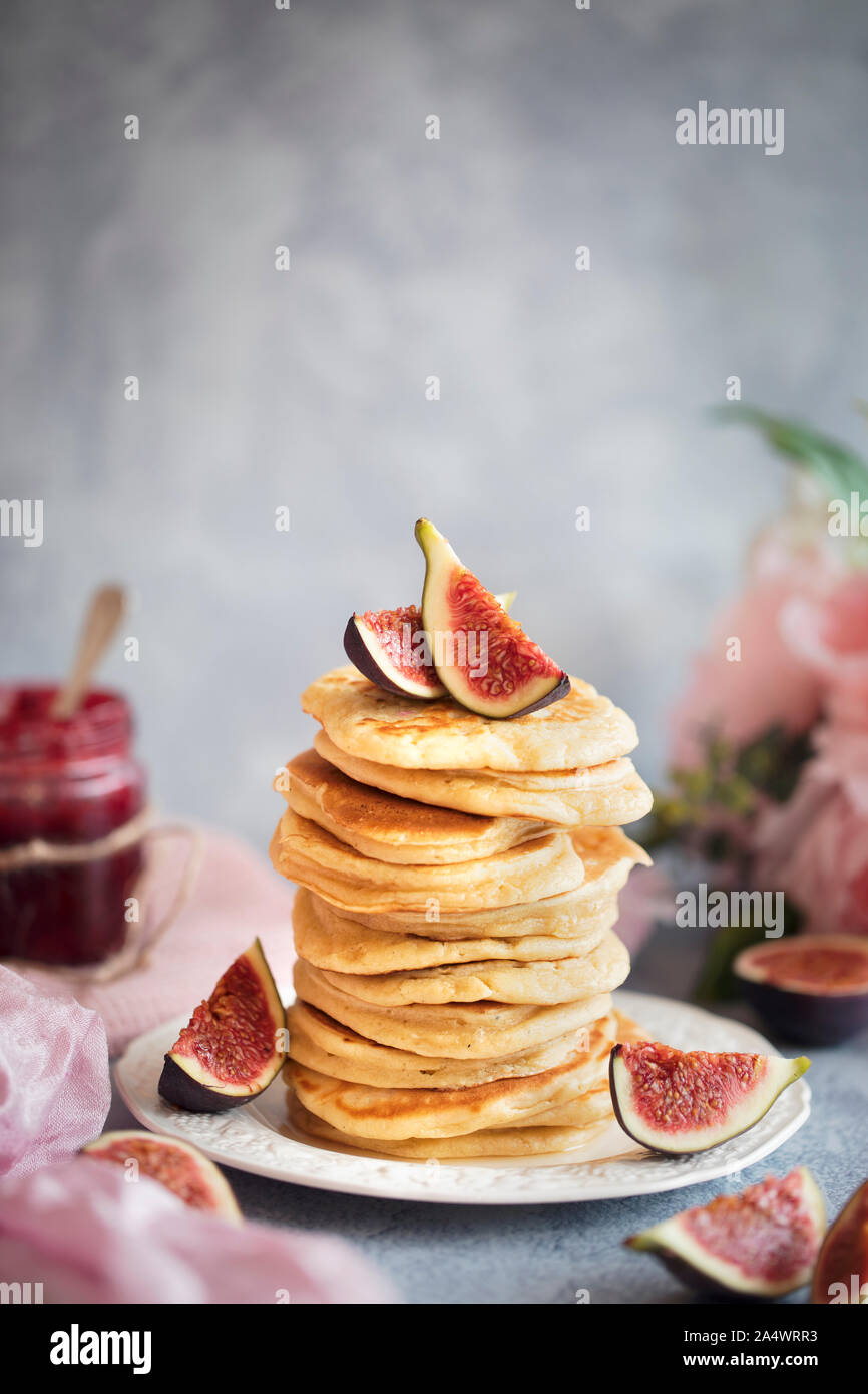 Una fresca prima colazione con una pila di frittelle e fichi freschi. Vi è un vasetto di marmellata in background, alcuni fiori e un pezzo di tessuto di colore rosa. Foto Stock