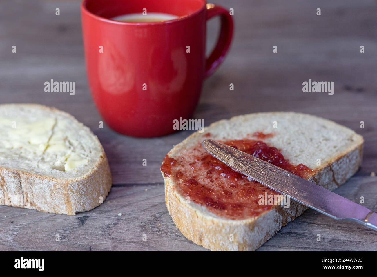 Un coltello fissa sulla parte superiore di una fetta di pane che ha jam sparsi su sulla parte superiore di una tavola di legno Foto Stock