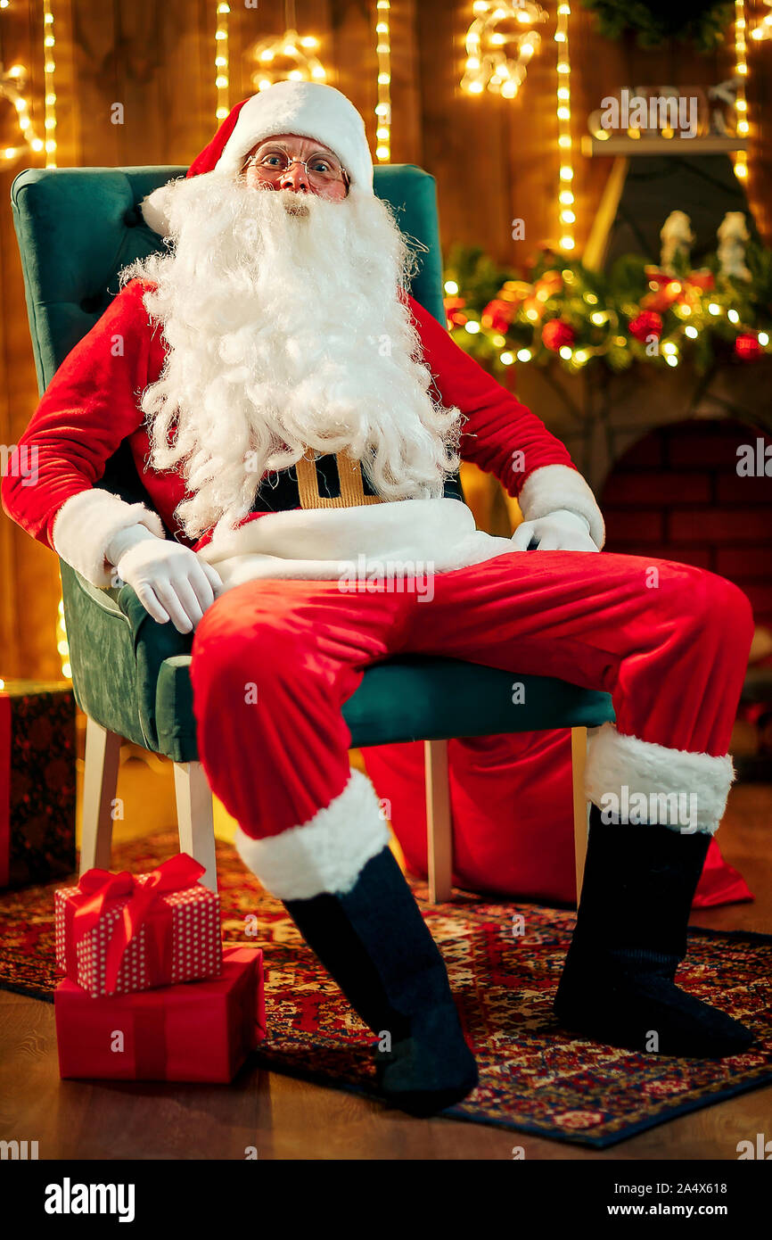 Foto di felice Santa Claus in occhiali seduti su una sedia e guardando la fotocamera Foto Stock