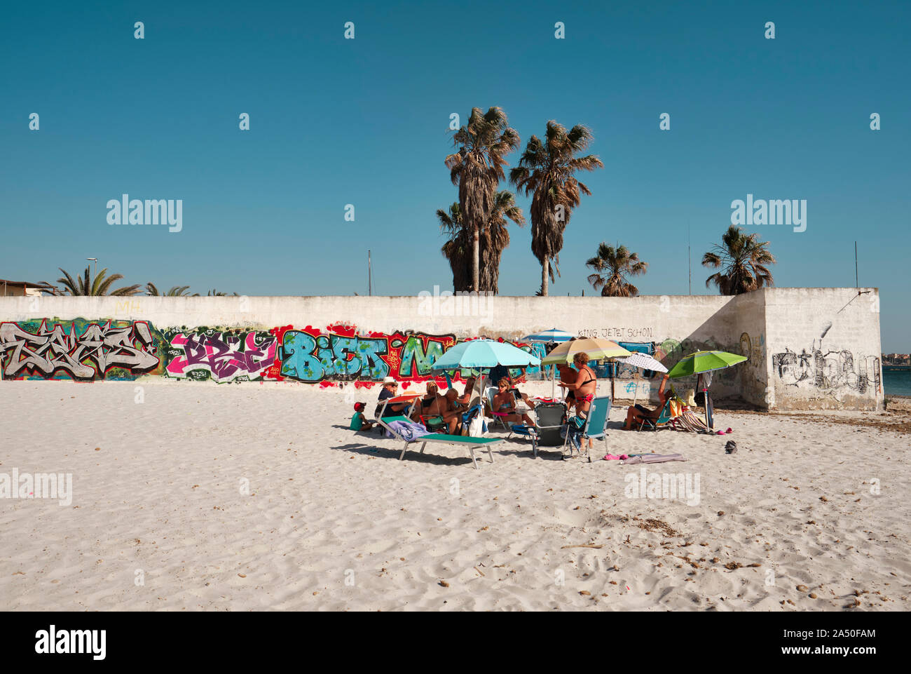 Famiglie e ombrelloni godendo la spiaggia sabbiosa a città di Alghero con un muro di graffiti in Sardegna Italia Europa Foto Stock