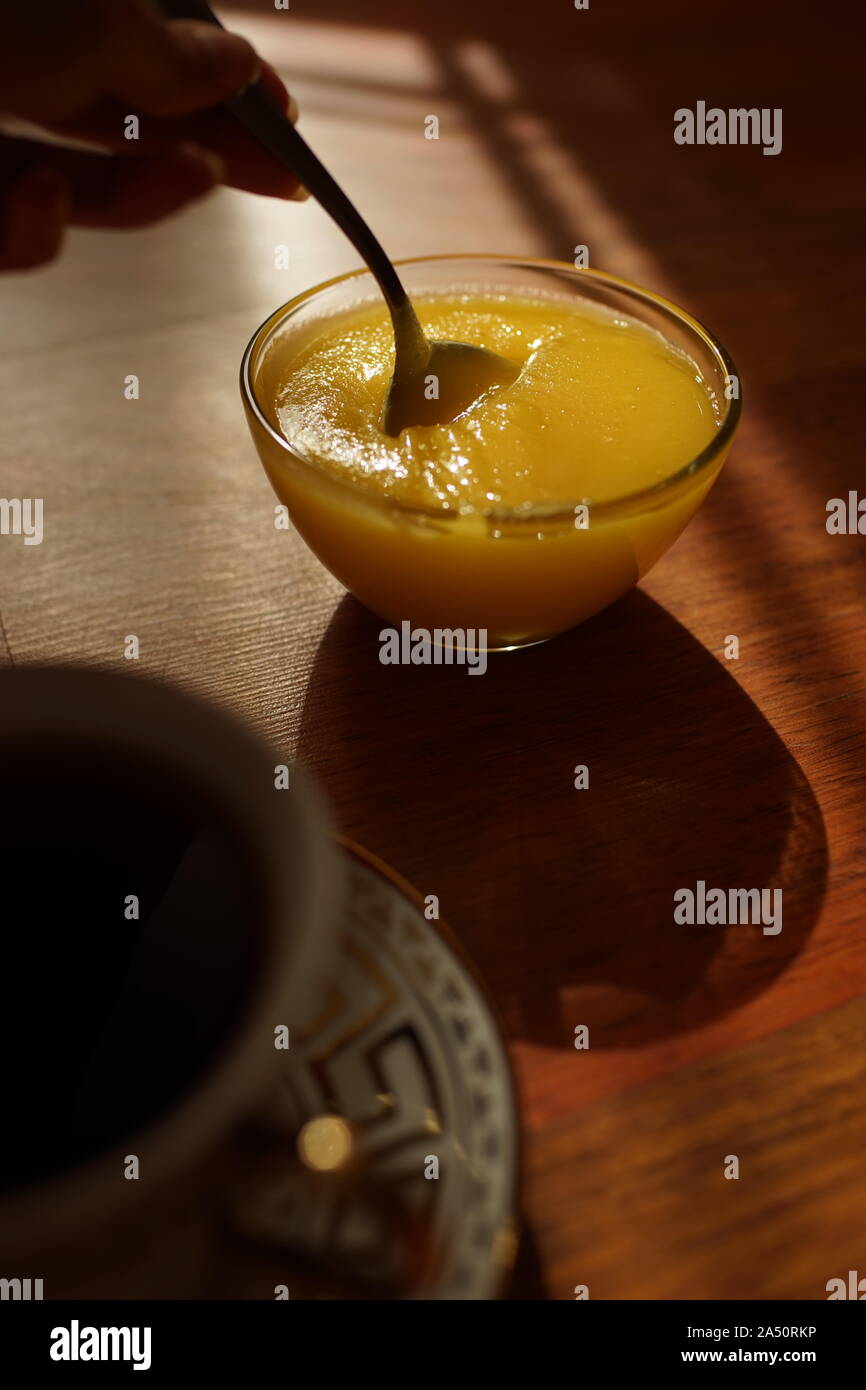 Round ciotola di vetro con il miele e il cucchiaio in mano su un soleggiato tavolo in legno Foto Stock