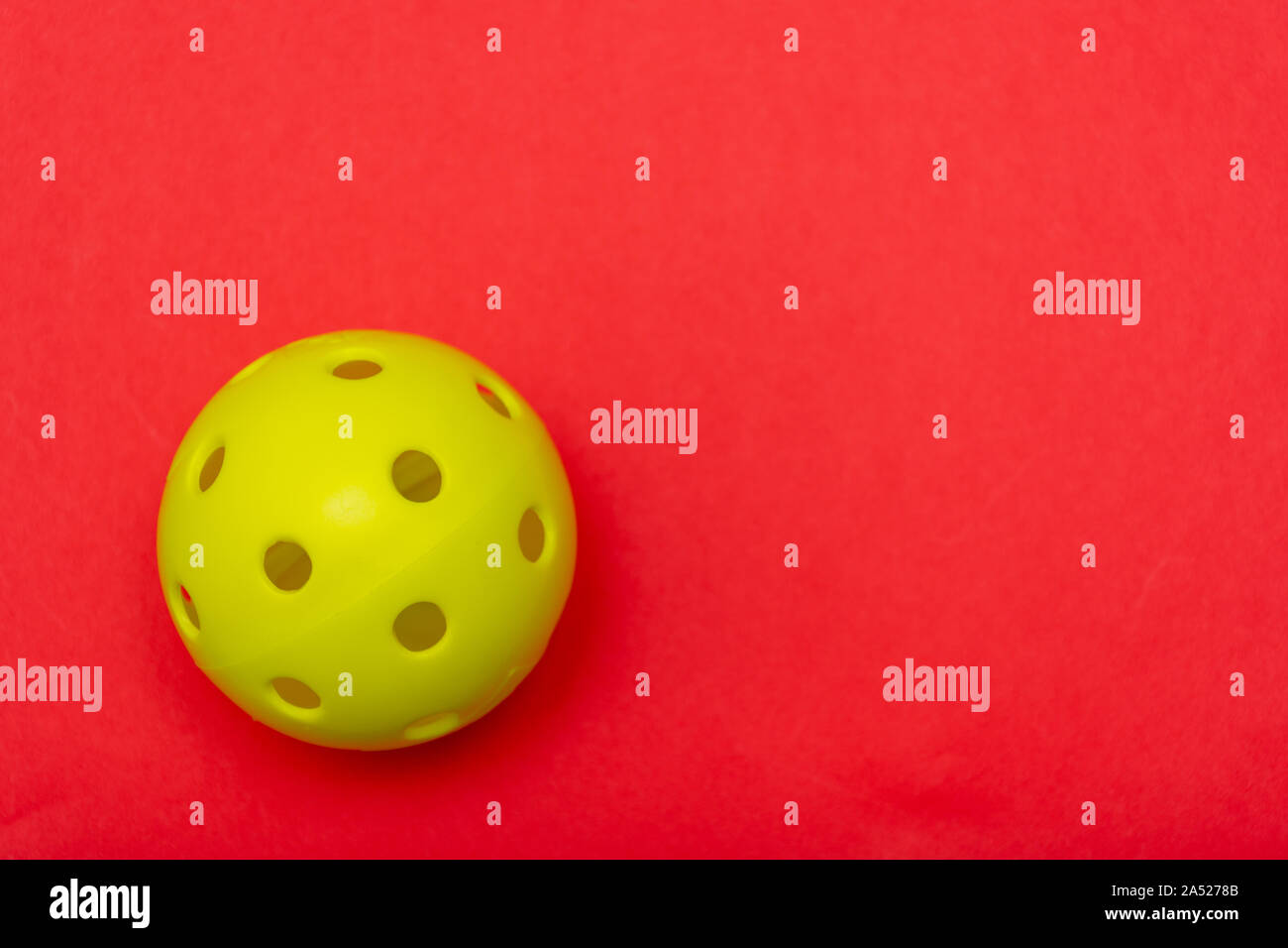 Di colore giallo brillante pickleball o sfera whiffle su un solido rosso luminoso appartamento sfondo laici che simboleggiano sport e attività con copia spazio. Foto Stock