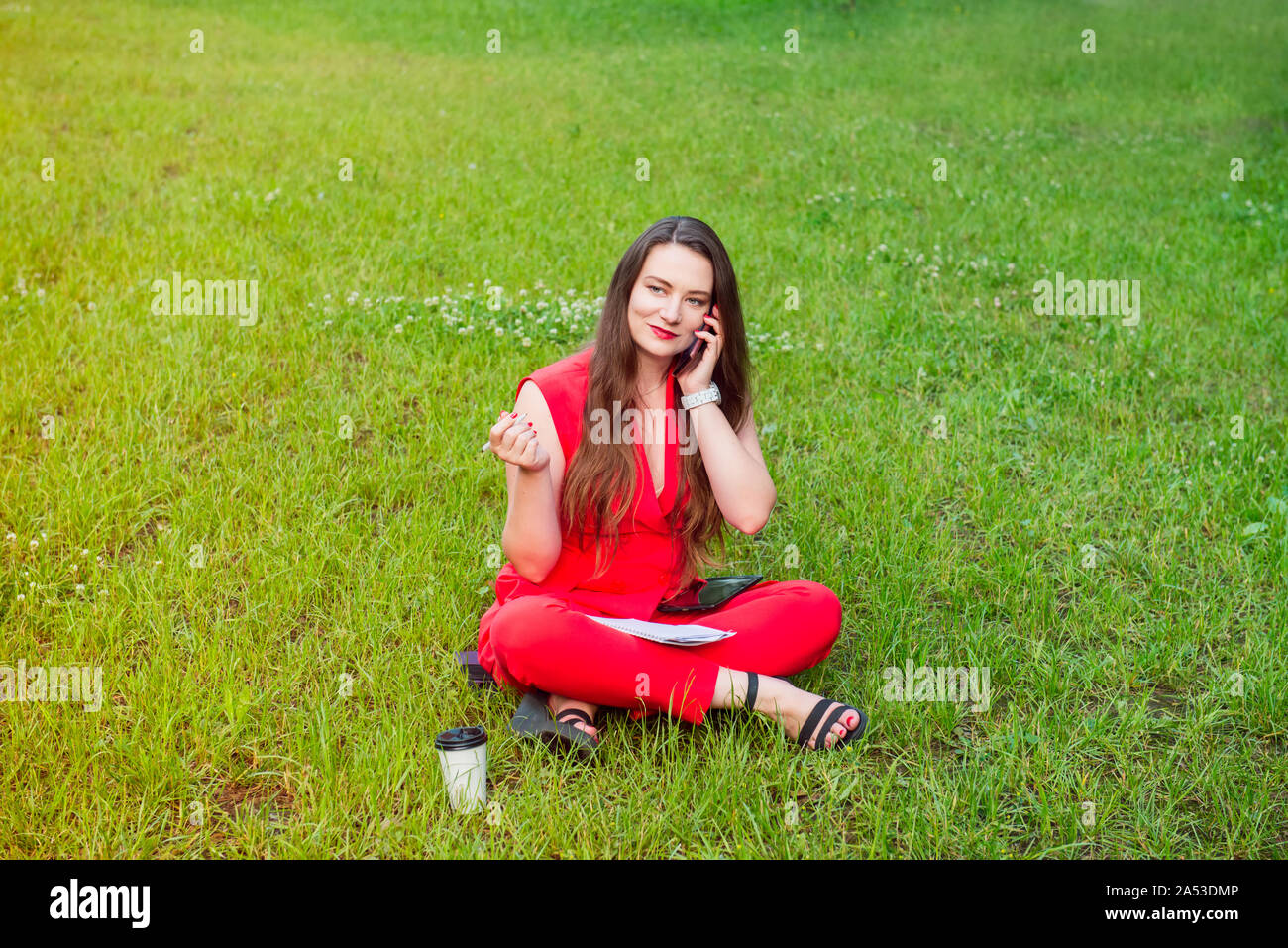 Giovane donna business plus dimensione in tuta rossa che parla il suo telefono e posti a sedere su uno sfondo di colore verde prato con notebook e penna in mano. ,Orking O Foto Stock